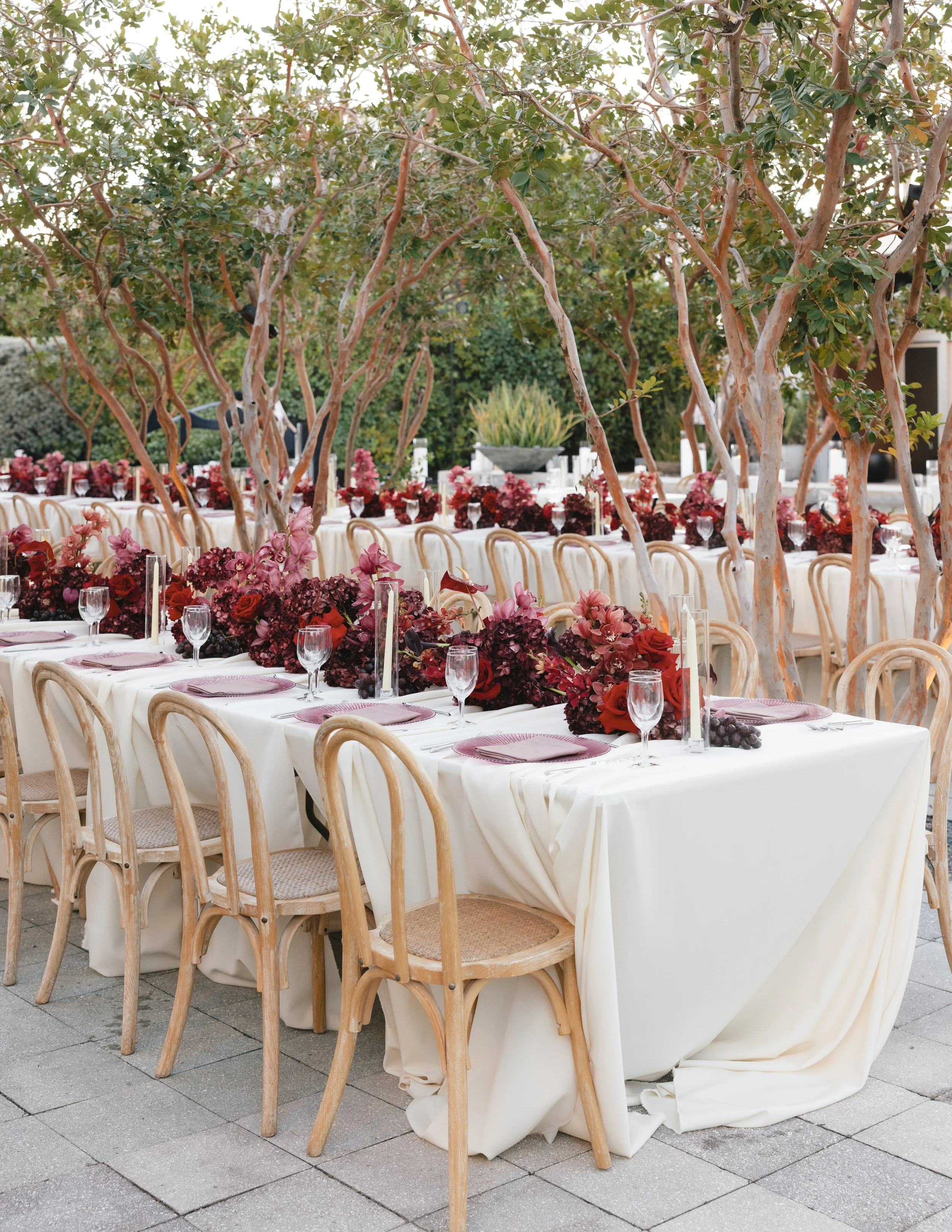 Outdoor event setup with long tables covered in white tablecloths, decorated with floral centerpieces, clear wine glasses, and pink napkins, surrounded by wooden chairs. Trees with twisting branches and green leaves shade the area, with additional fl