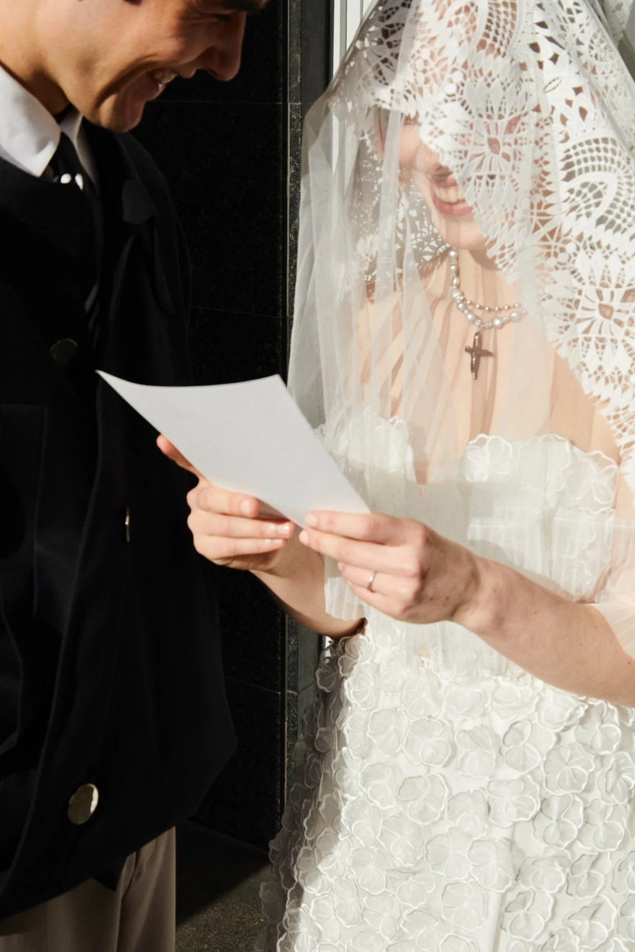 A bride and groom are smiling and looking at a piece of paper, with the bride wearing a lace veil and a pearl necklace with a cross pendant.