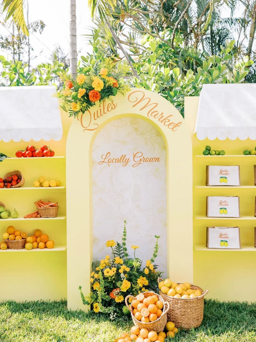 Market stand with fresh fruits, yellow flowers, and a sign labeled 'Quiles Market, Locally Grown' amid greenery.