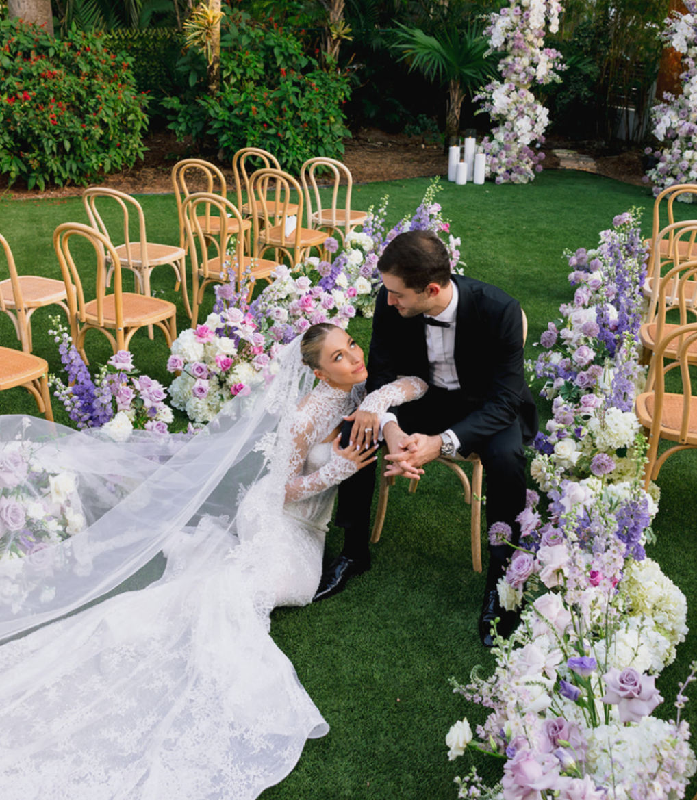 Bride and groom sitting on a chair surrounded by purple and white flowers at an outdoor wedding ceremony.