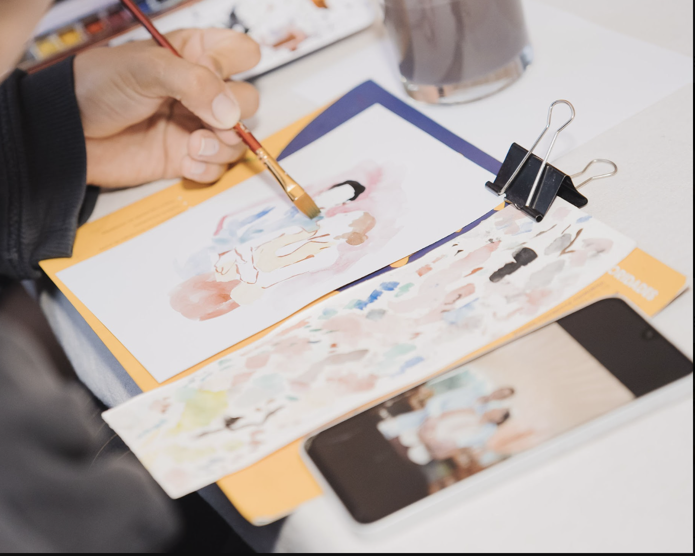 Person painting watercolor art on paper at a desk with a smartphone, paint palette, and binder clip.
