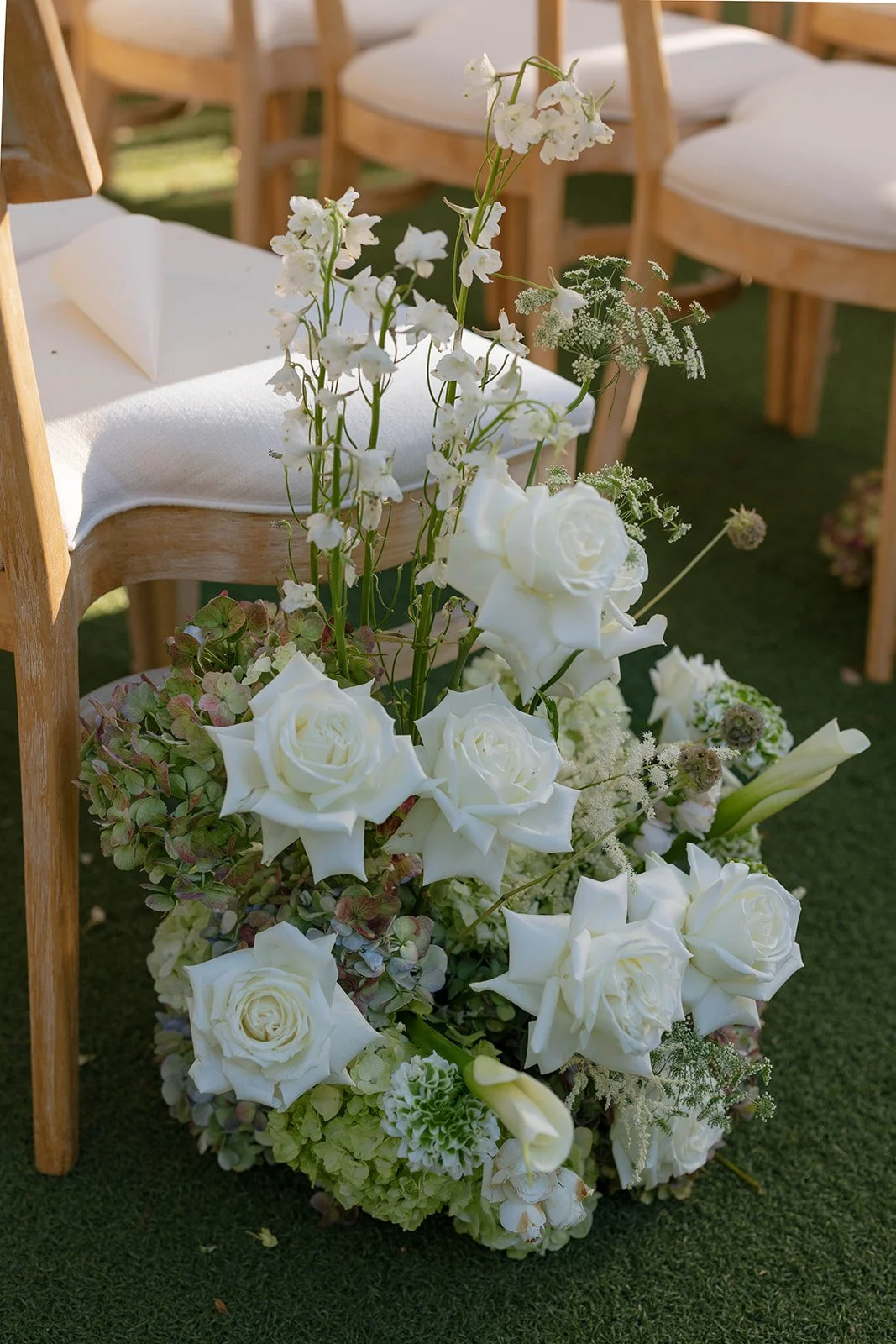 A floral arrangement of white roses, white calla lilies, and other white flowers, placed beside light wooden chairs with white cushions on green grass.