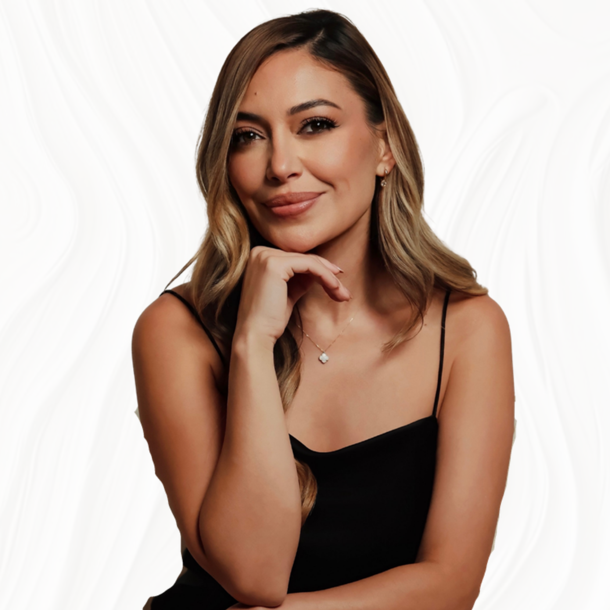 A woman posing with her chin resting on her hand, wearing a black spaghetti strap top and jewelry, and smiling at the camera against a plain white background.