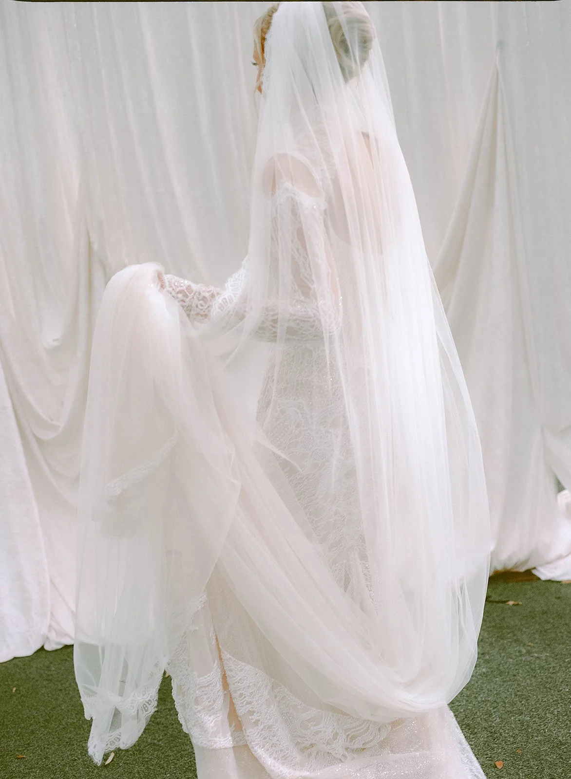 A bride in a white wedding gown with lace details, lost in thought and holding her gown's train under a sheer veil.