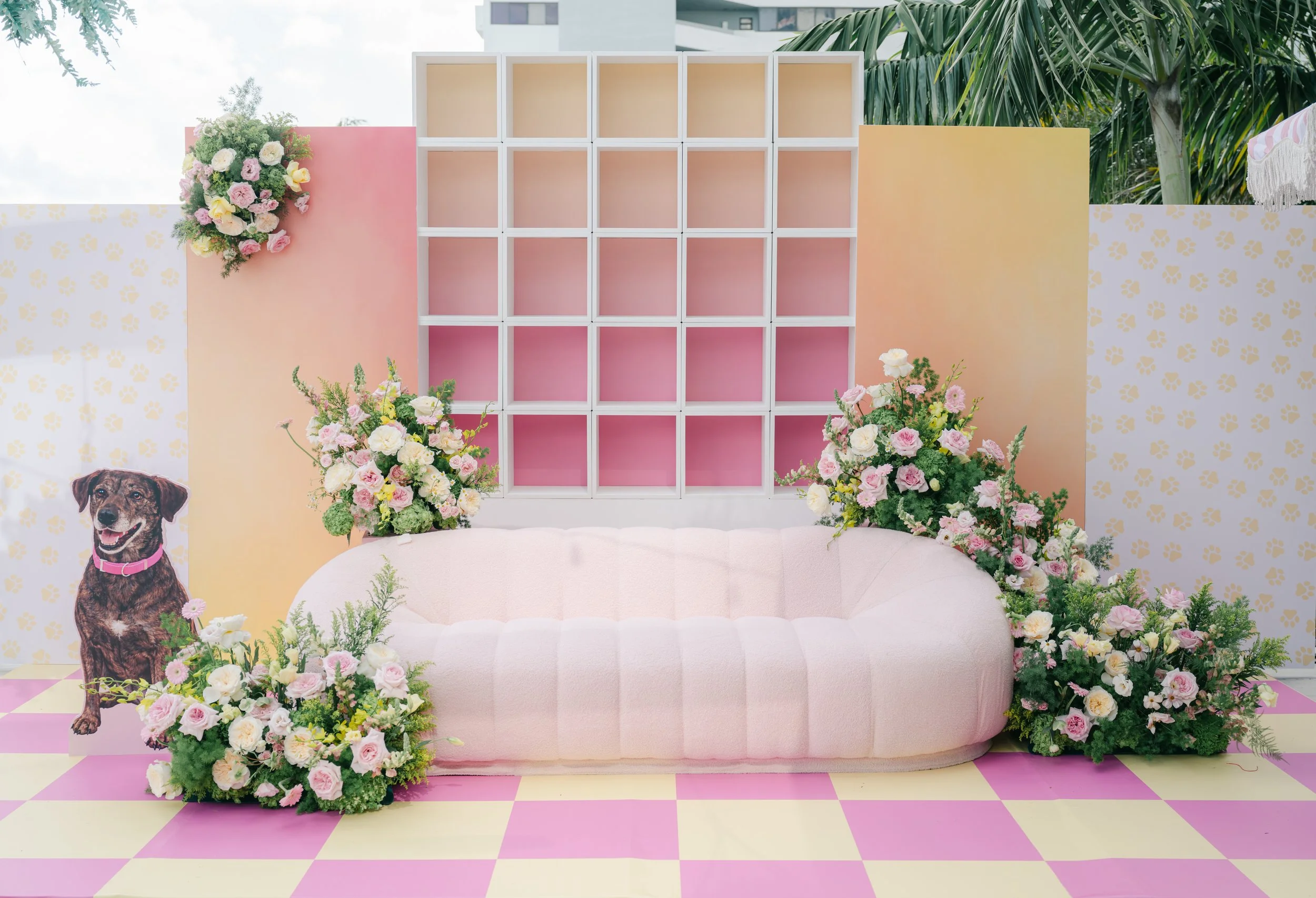 Decorative backdrop with pink, yellow, and white panels, floral arrangements, a pink sofa, a dog image, and a checkered pink and yellow floor.