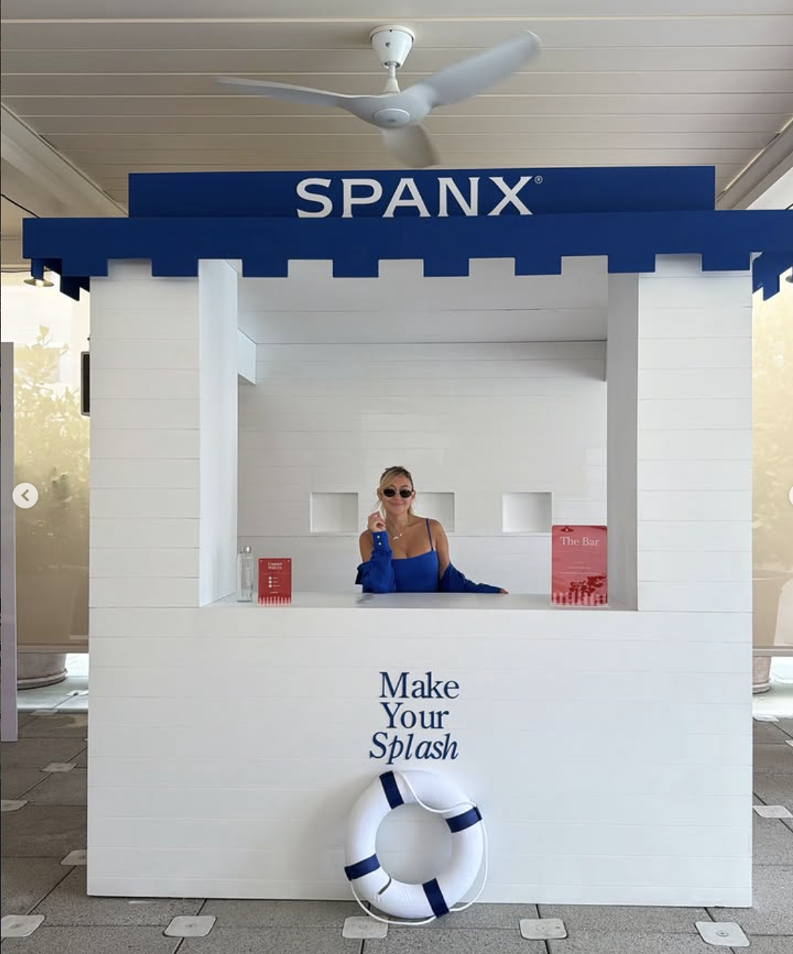 A woman in sunglasses wearing a blue top standing behind a white beach-themed booth with the sign 'SPANX.' The booth encourages visitors to 'Make Your Splash' and features a lifebuoy hanging at the bottom. The background includes an indoor ceiling fa