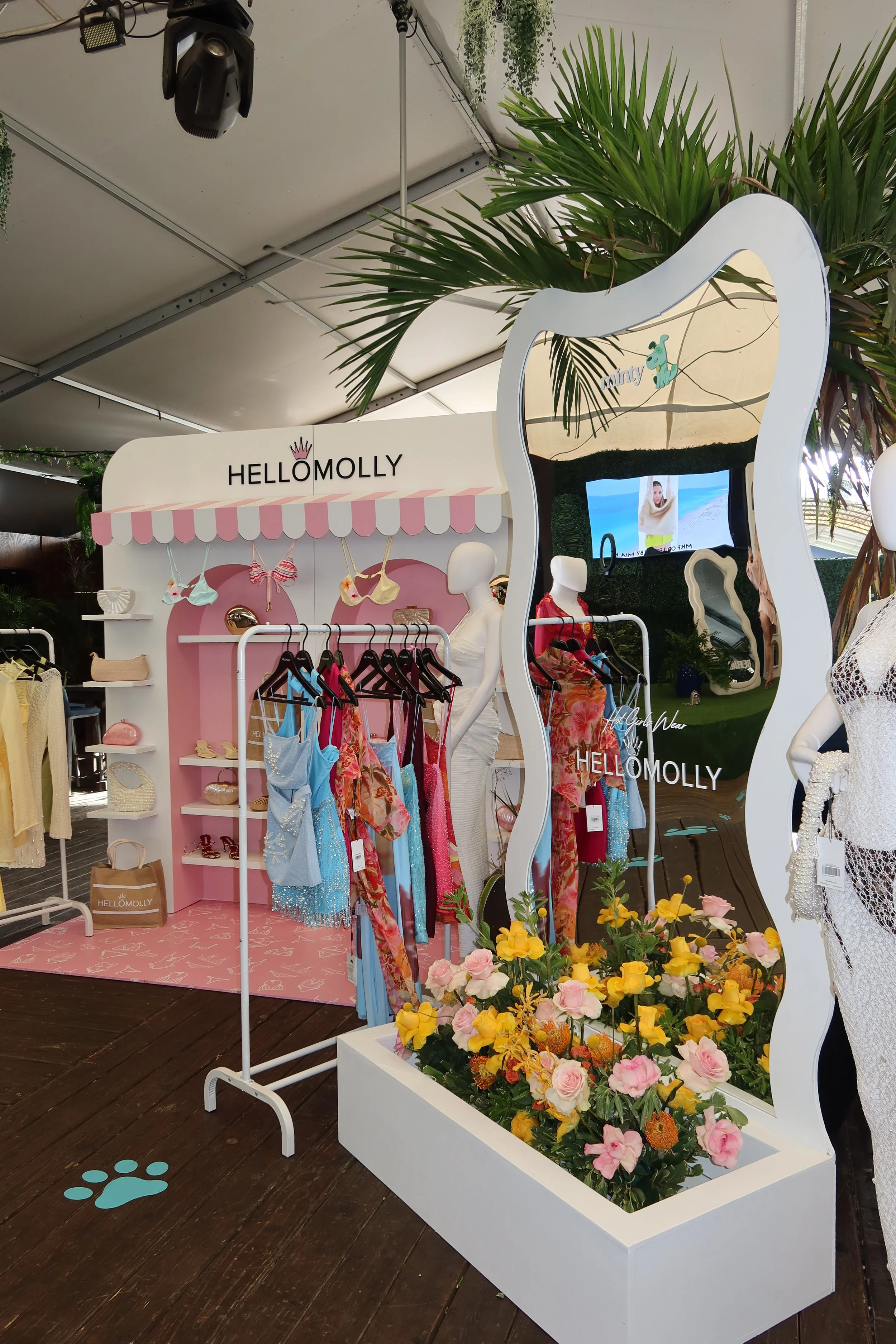 A boutique display booth with the name 'Hello Molly' featuring clothing, accessories, and mannequins, decorated with flowers and tropical palm trees.