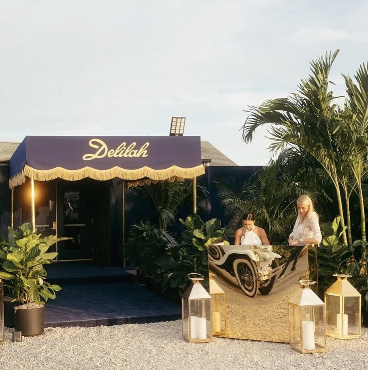 A garden scene outside a building with a purple awning labeled "Delilah" in cursive. Two women are standing behind a large reflective table, with one wearing a white top and the other a sleeveless white dress. There are decorative lanterns and candle