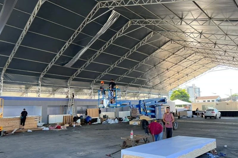 Construction workers installing a large fabric roof in an indoor venue with a concrete floor and some cars parked outside.