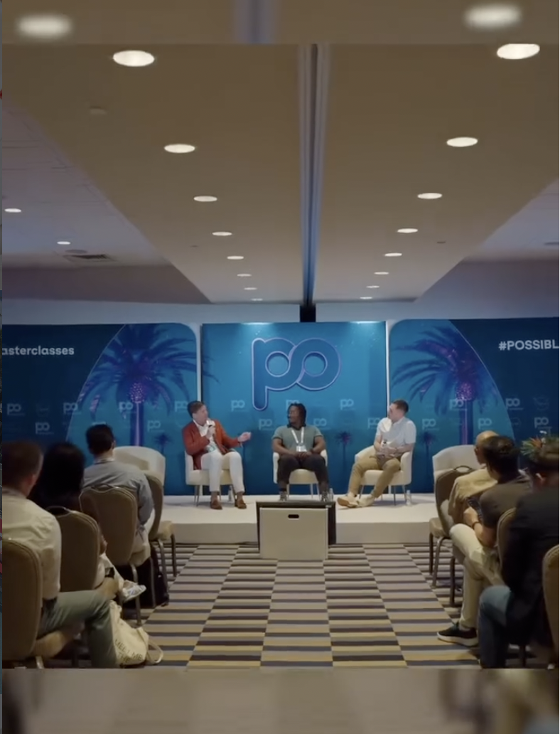 Three men sitting on a stage in front of an audience at a conference or panel discussion with a blue backdrop featuring the letters 'po' and tropical palm tree designs.