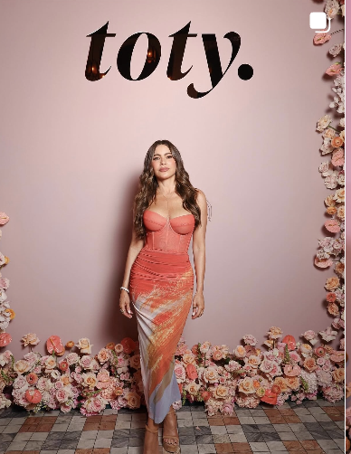 Woman in a pinkish-orange dress standing in front of a pink wall with the word "tory." written above her, surrounded by floral decorations at the bottom.