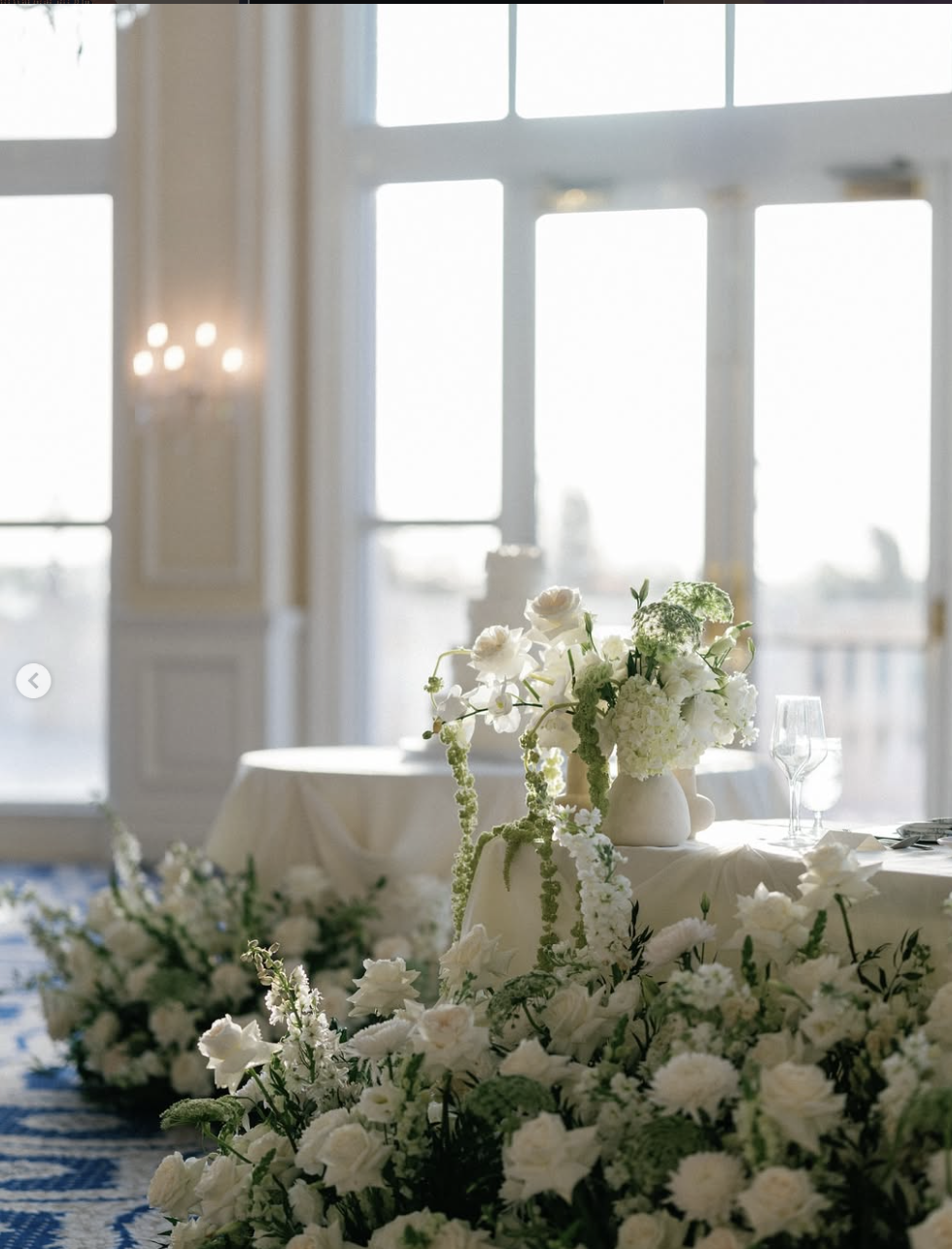 A banquet table decorated with a white cloth, adorned with a large floral arrangement of white flowers, and set with wine glasses and silverware, in front of large windows letting in natural light.