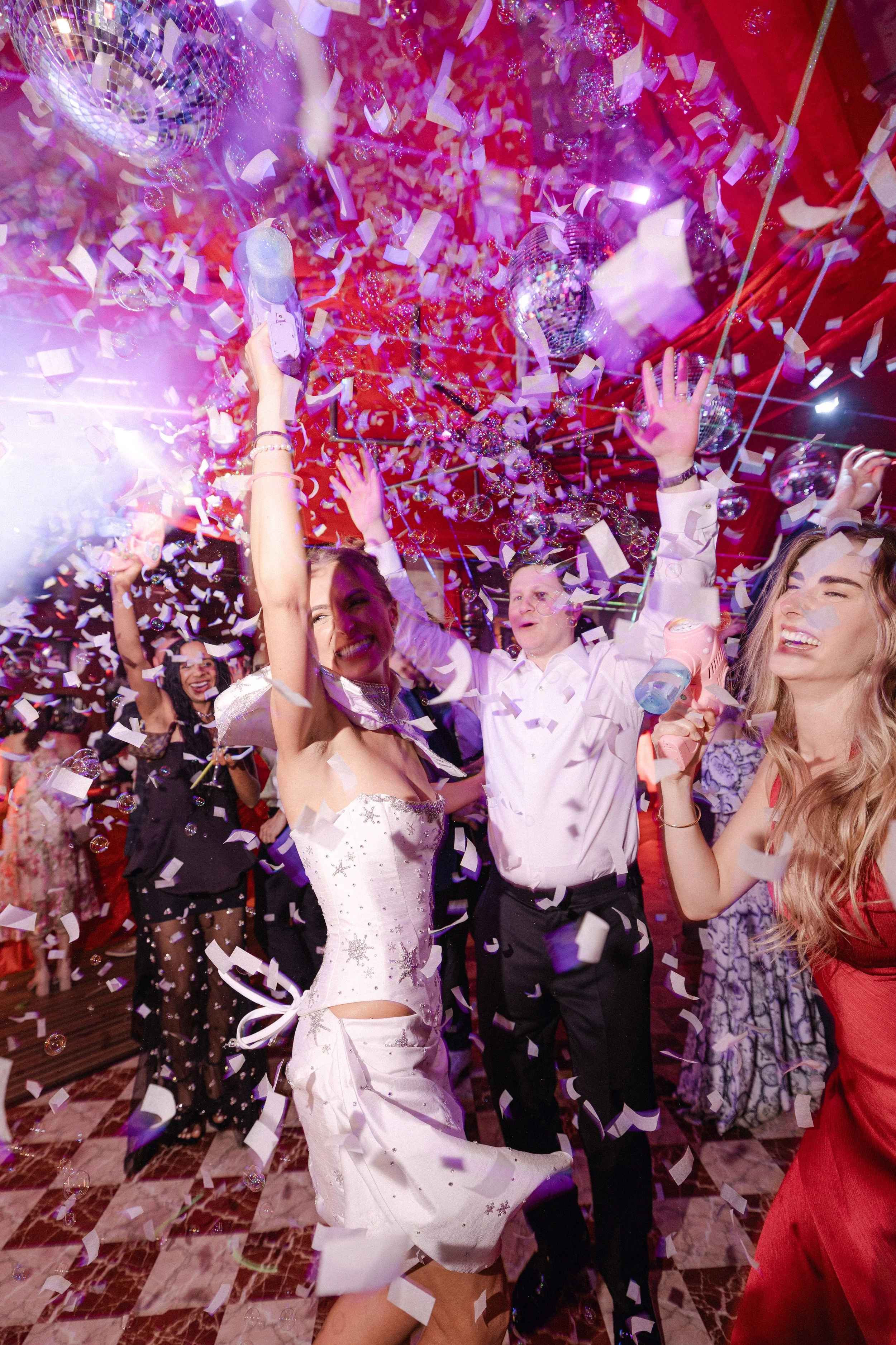 People dancing and celebrating at a party with confetti, balloons, and a disco ball.
