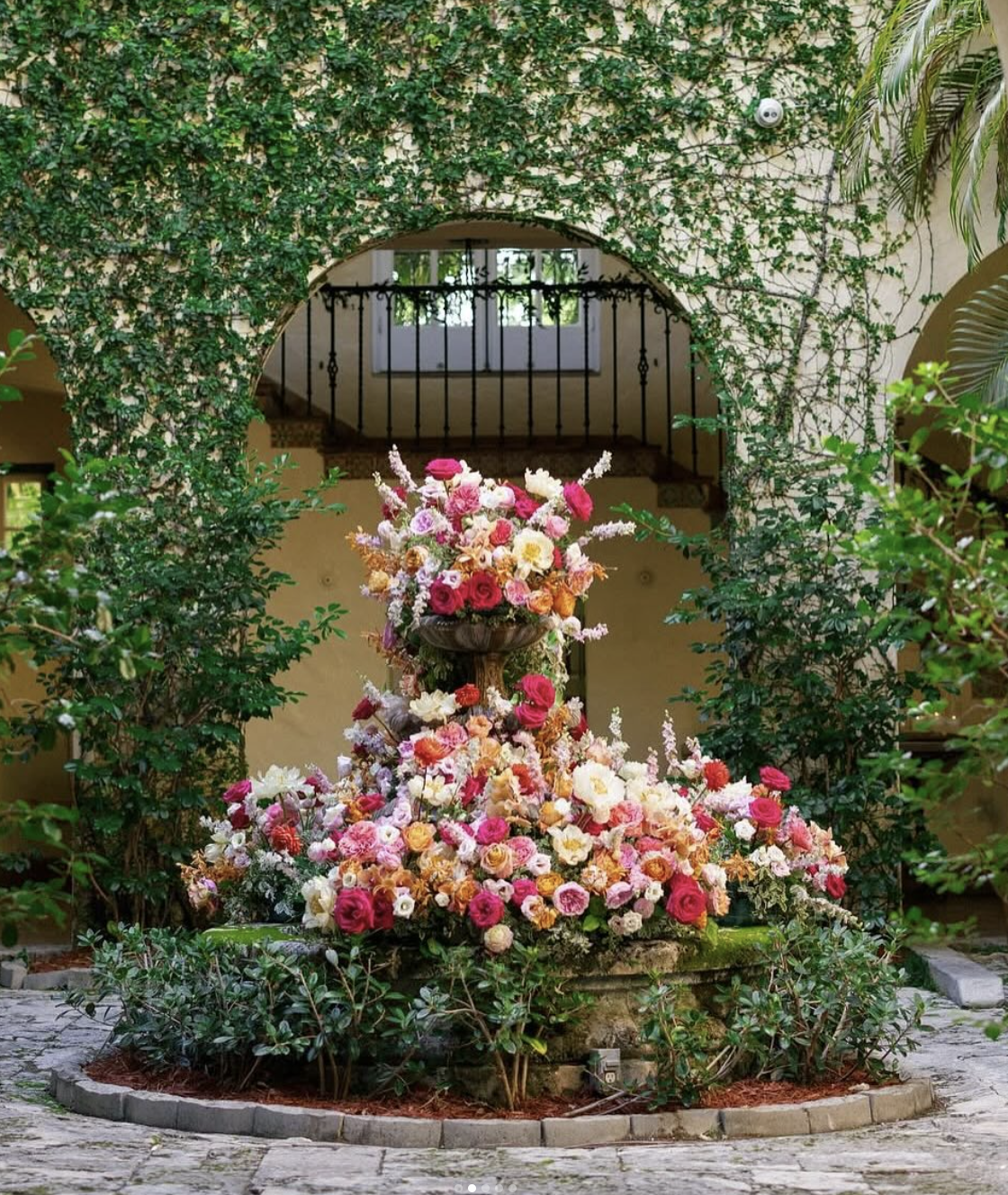 Arrangement of pink, white, orange, and red flowers in a stone fountain surrounded by green foliage inside a building.