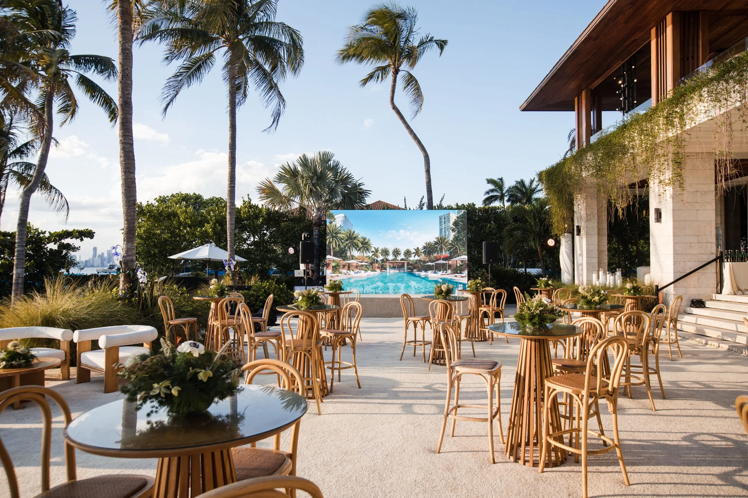 Outdoor event space with wooden chairs and round tables decorated with flowers, overlooking a swimming pool, surrounded by palm trees and modern buildings.