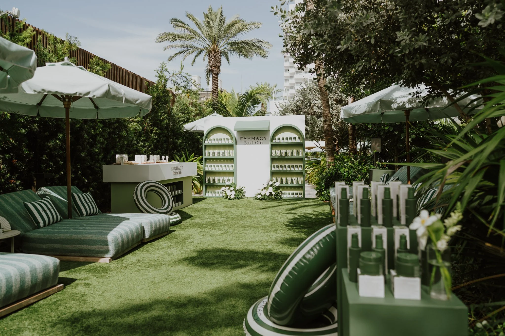 Outdoor setting with green striped cushions on lounge chairs, white umbrellas, a backdrop of lush greenery including palms, and white shelves with skincare products at a beach club.
