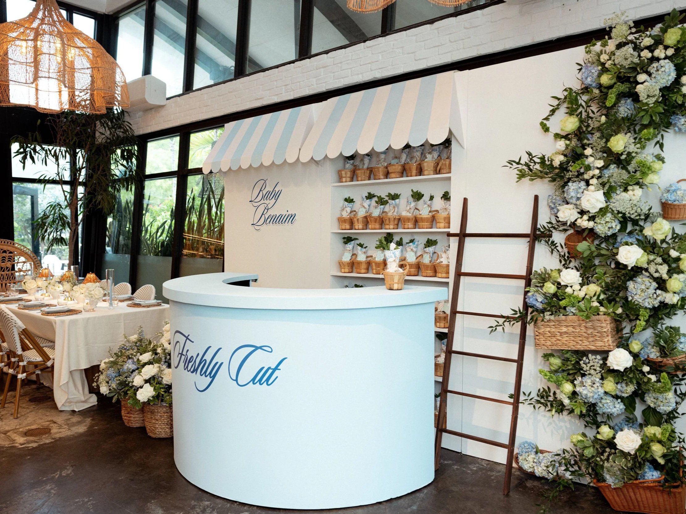 Decorative event setup with floral arrangements, a white booth labeled "Baby Benaim," and a table with place settings, inside a modern venue with large windows.