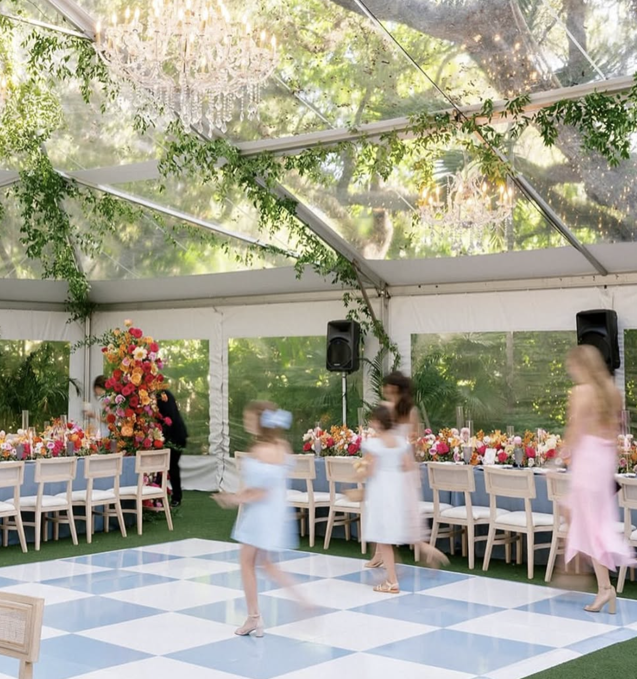 A decorated event tent with a ceiling chandelier, green foliage, and floral centerpieces on tables. Several people are dancing on a checkered dance floor inside, with some people blurred in motion.