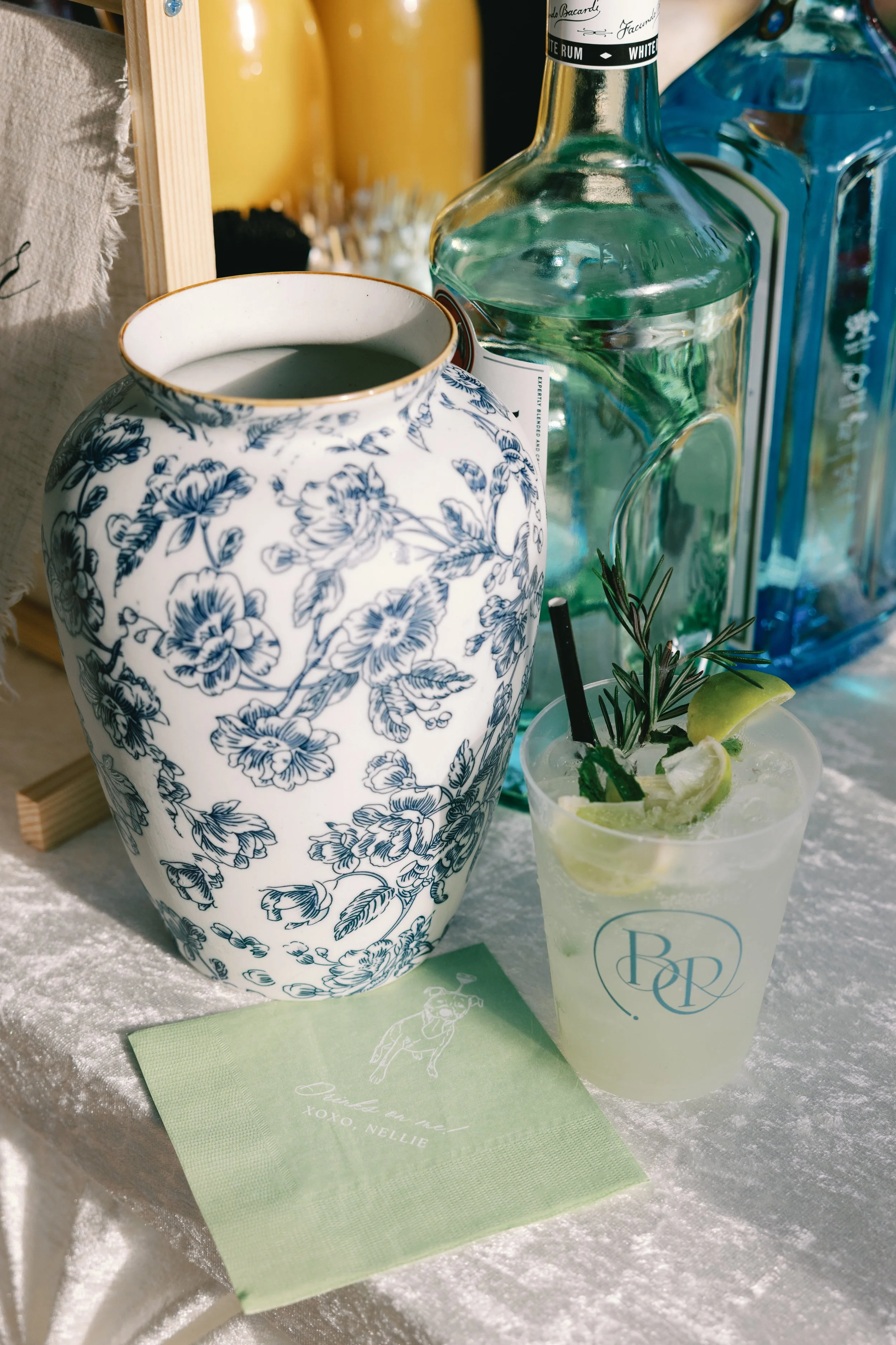 A white ceramic vase with blue floral patterns, a clear glass bottle of white rum, a glass with a cocktail garnished with lemon, lime, and rosemary, and a green paper napkin on a textured white table.