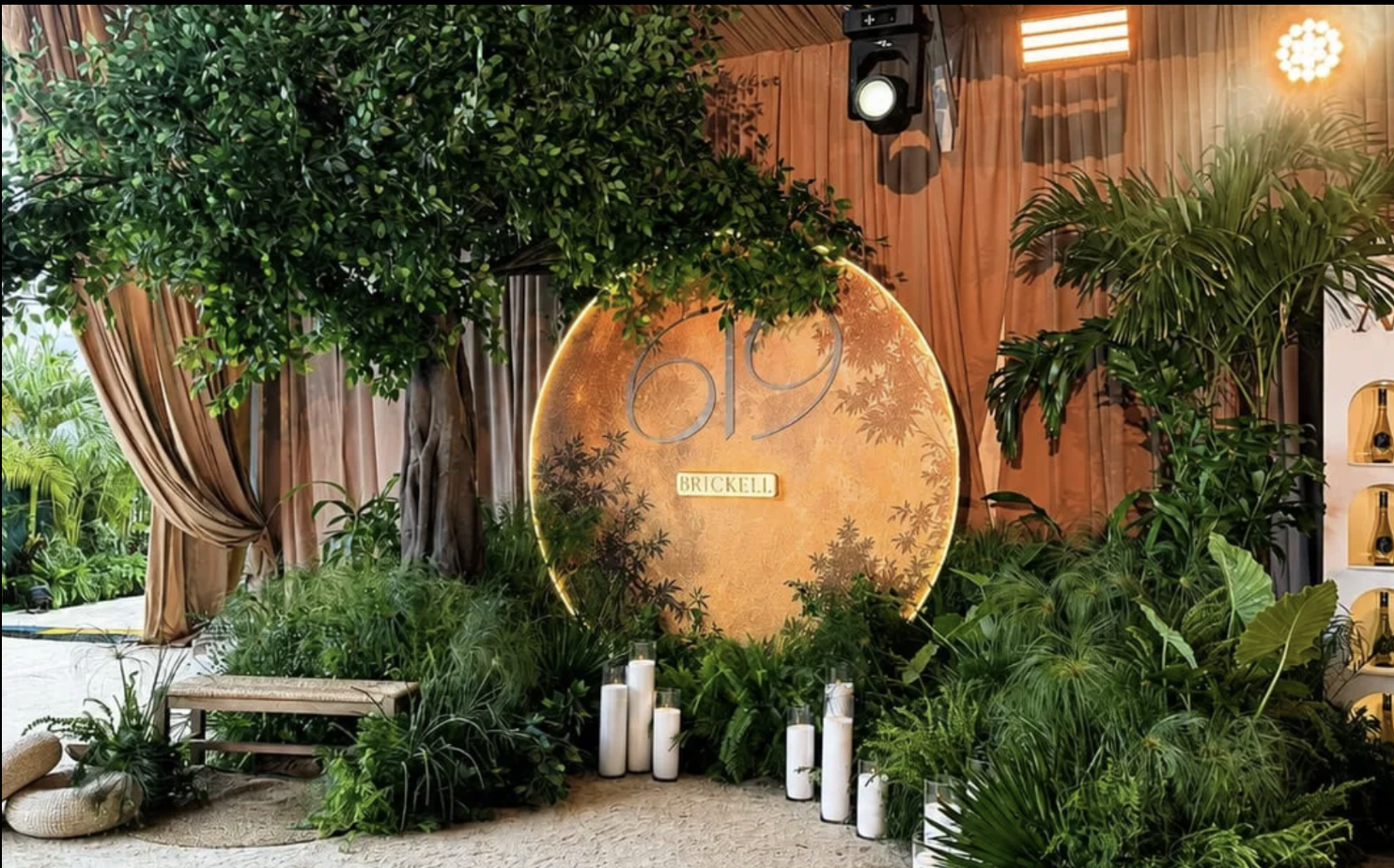 Indoor event setup with lush green plants and trees surrounding a circular, illuminated sign that reads '19' and 'BRICKELL.' Candles are placed on the floor in front of the sign, and orange curtains hang in the background with stage lights overhead.