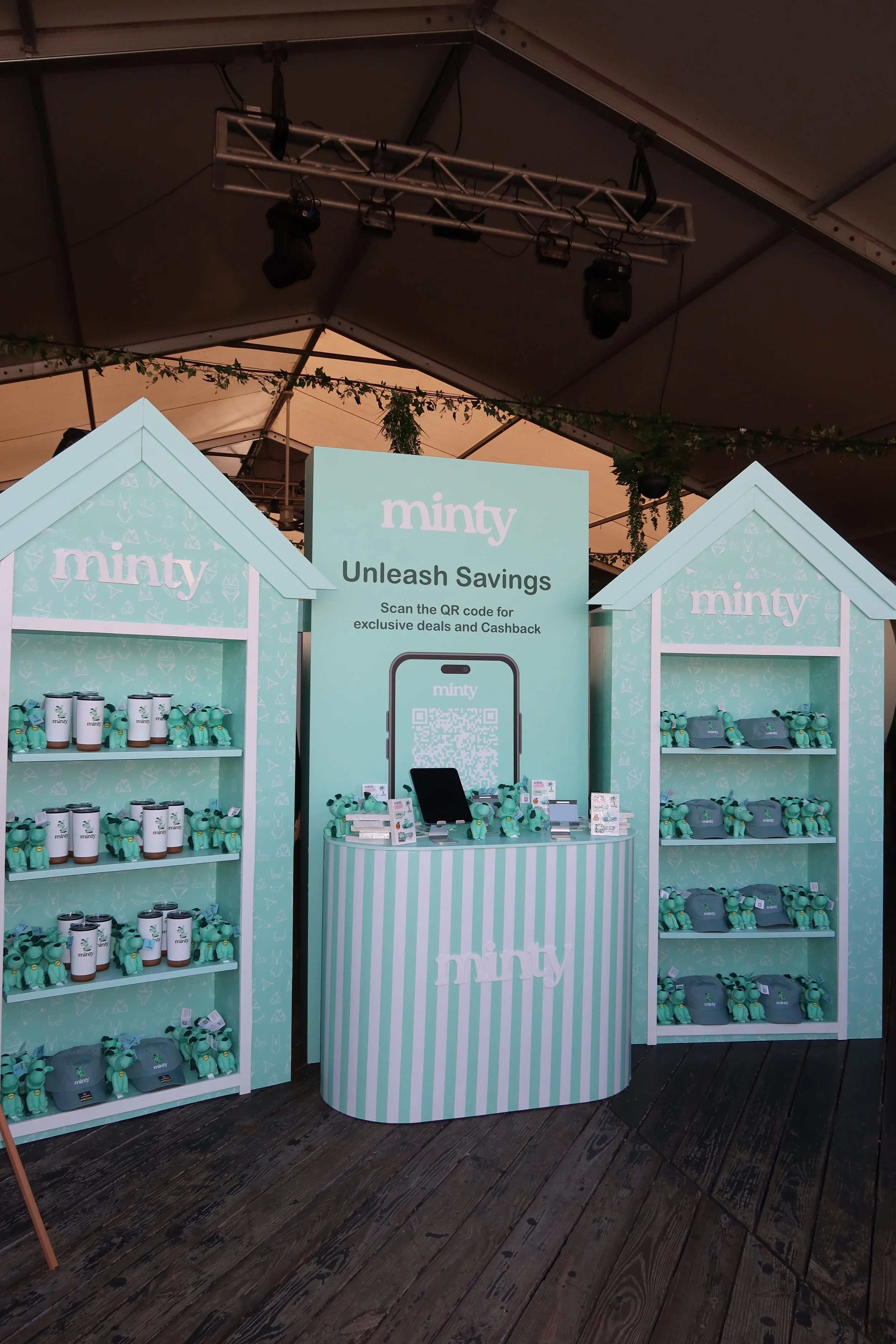 A display booth for Minty products with shelves of cups, hats, and plush toys, and a central stand with a QR code for discounts, set inside a tent with decorative greenery and lighting.