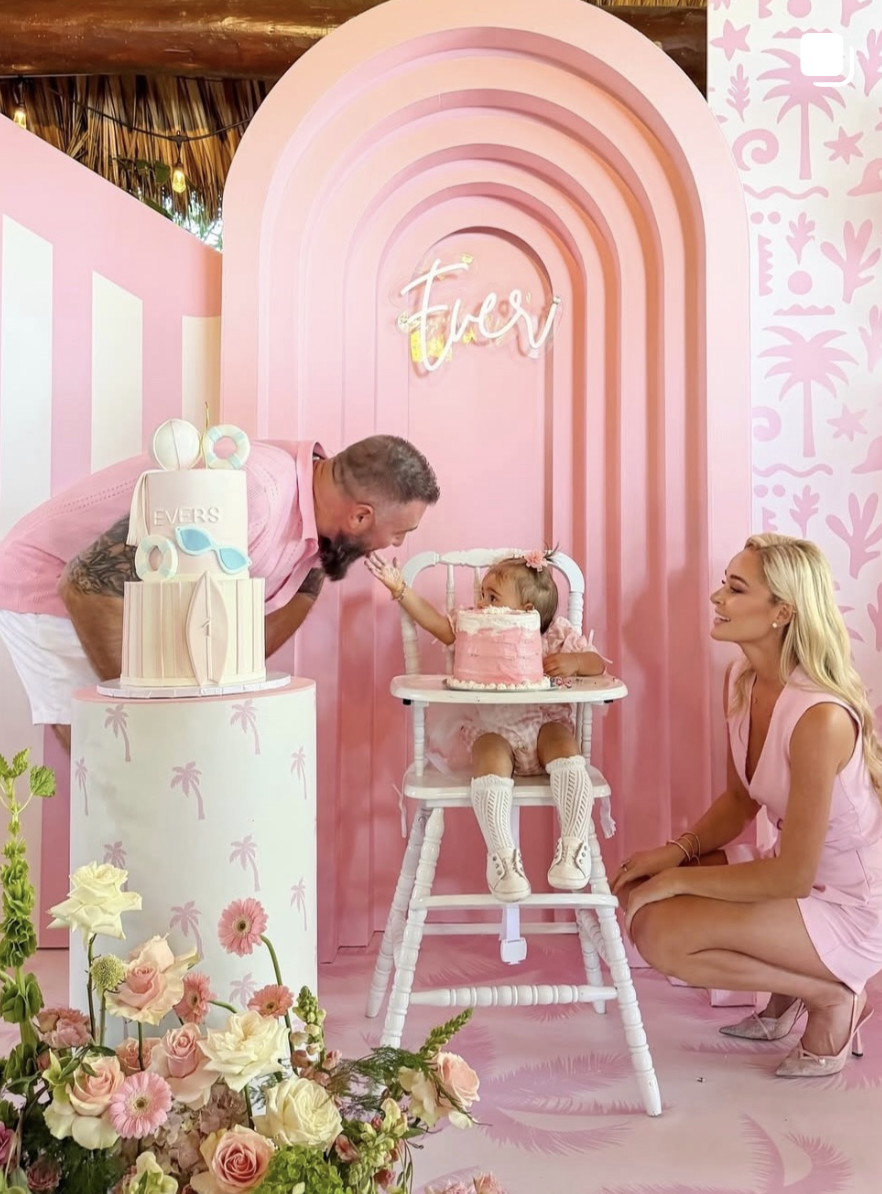 A family celebrating a birthday in a pink-themed setting with flowers, a cake, and a birthday sign that says 'Evers'. The little girl, seated in a high chair, reaches for the man leaning towards her. The woman kneeling beside her is smiling.