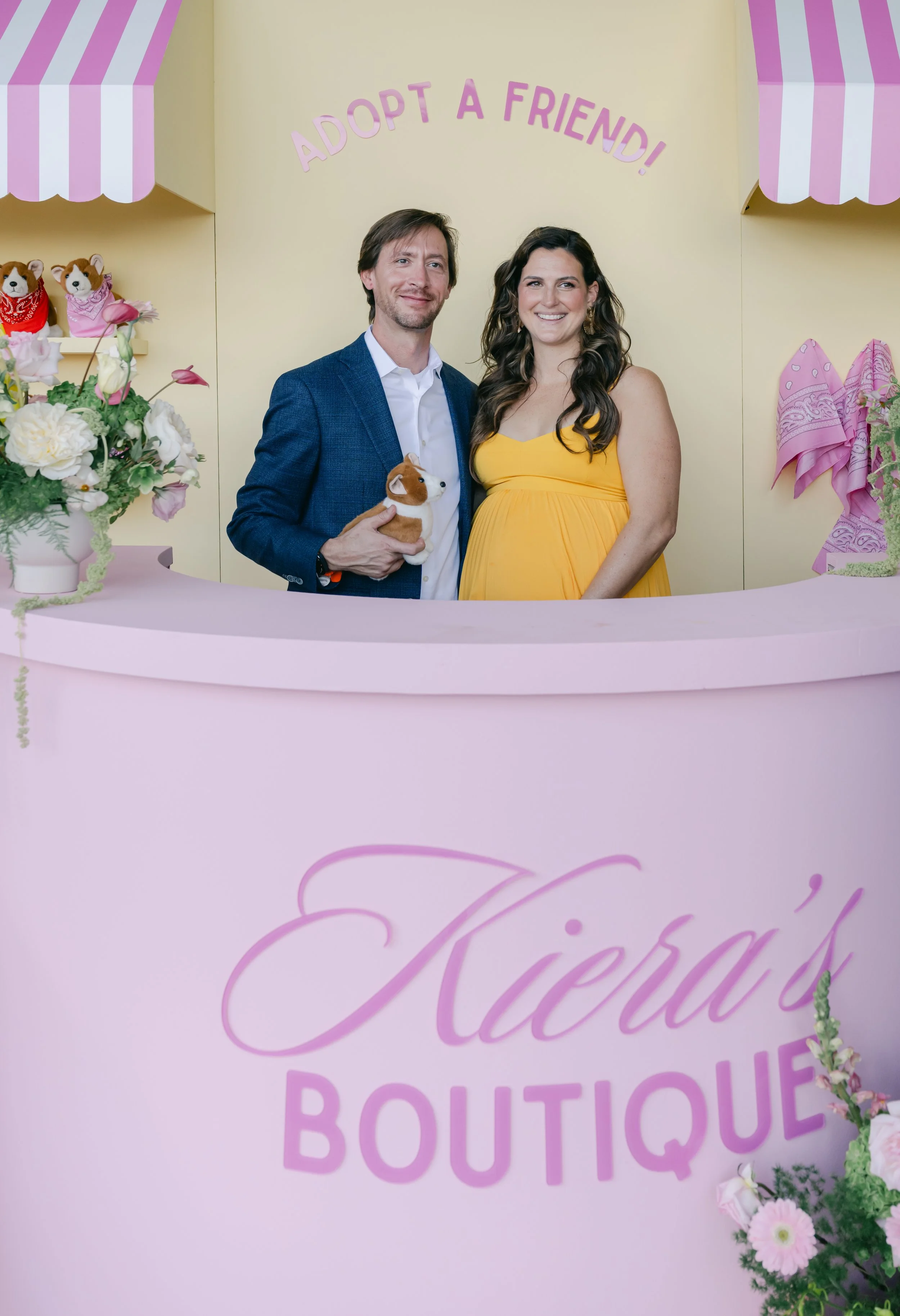A man and a woman smiling at a pink boutique counter with flowers and stuffed animals, with a sign that says "ADOPT A FRIEND!" in the background.