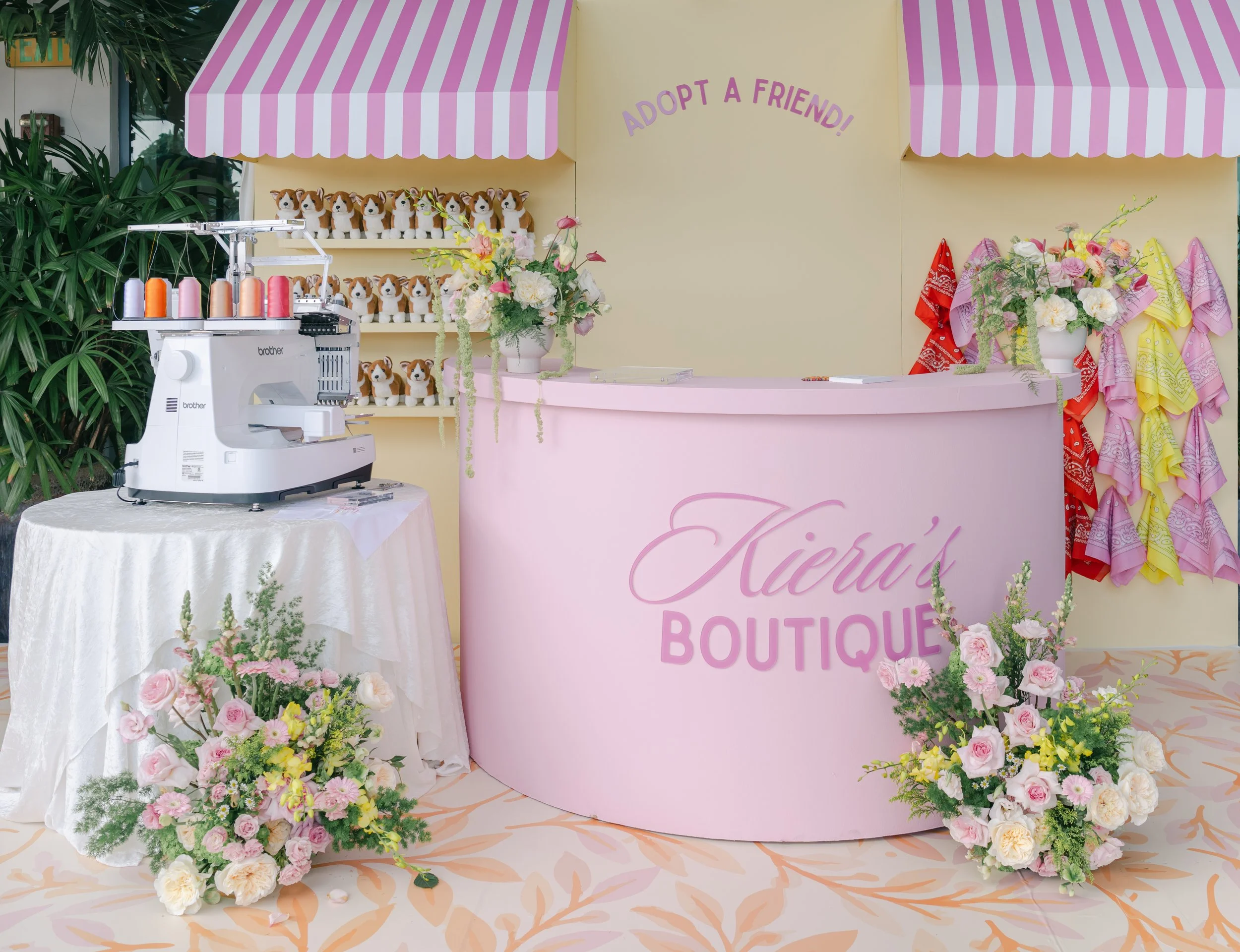 A pink and white booth with artificial dog plush toys on shelves, colorful spools of thread, and flowers, with a sign that says 'Adopt a Friend!' and 'Kiera's Boutique'.
