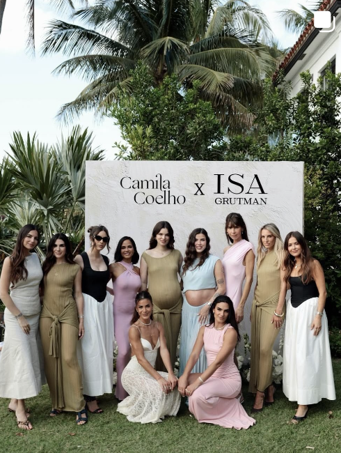 Group of ten women posing outdoors in front of a white backdrop with text that reads "Camila Coelho x ISA Grutman." Tall palm trees and lush green foliage are in the background.
