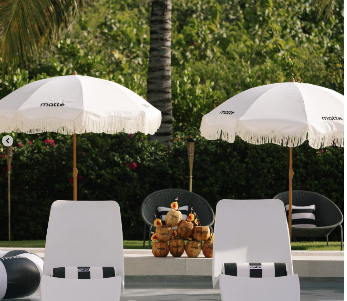 Poolside lounge area with two white umbrellas, white lounge chairs, a stack of coconuts with small drink umbrellas, and tropical greenery in the background.
