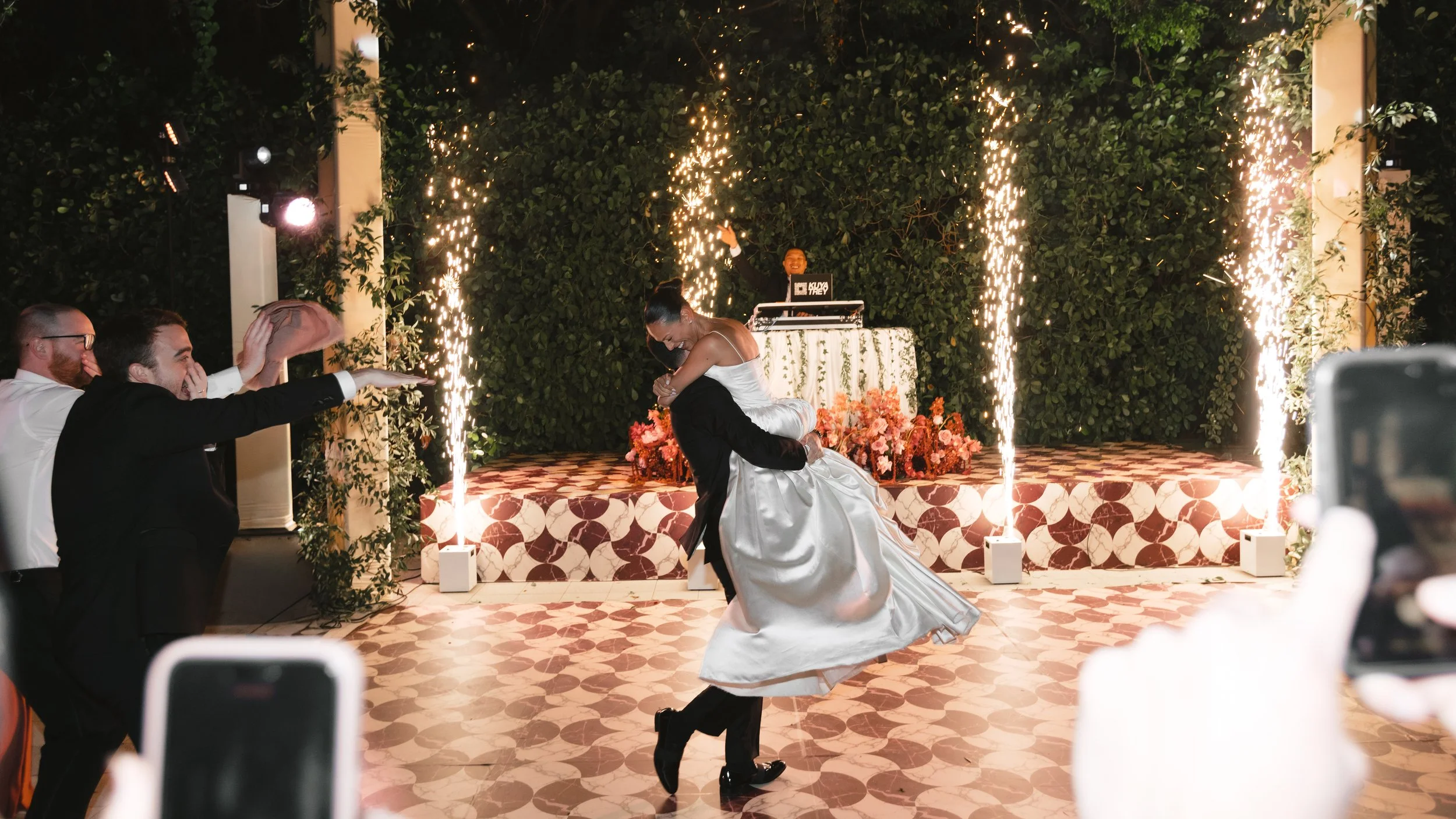 A newlywed couple performing their first dance at a wedding reception, surrounded by guests, fireworks, and a DJ booth in the background.