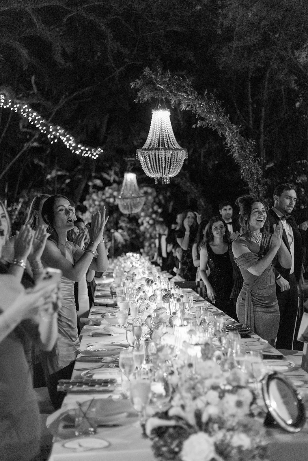 Black and white photo of a formal dinner party with people standing and clapping around a long, elegantly decorated table, hanging chandeliers, and a lush outdoor setting at night.