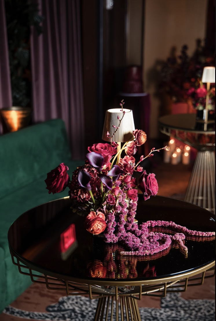 A black round table with a floral centerpiece, a small lamp, and pink beaded necklace. The background has a green sofa, curtains, and some lit candles.