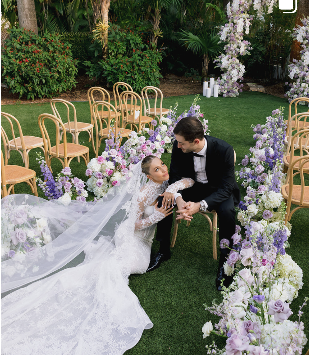 A bride and groom sitting on the grass at their wedding, surrounded by purple and white flowers, with empty chairs around them in an outdoor garden setting.