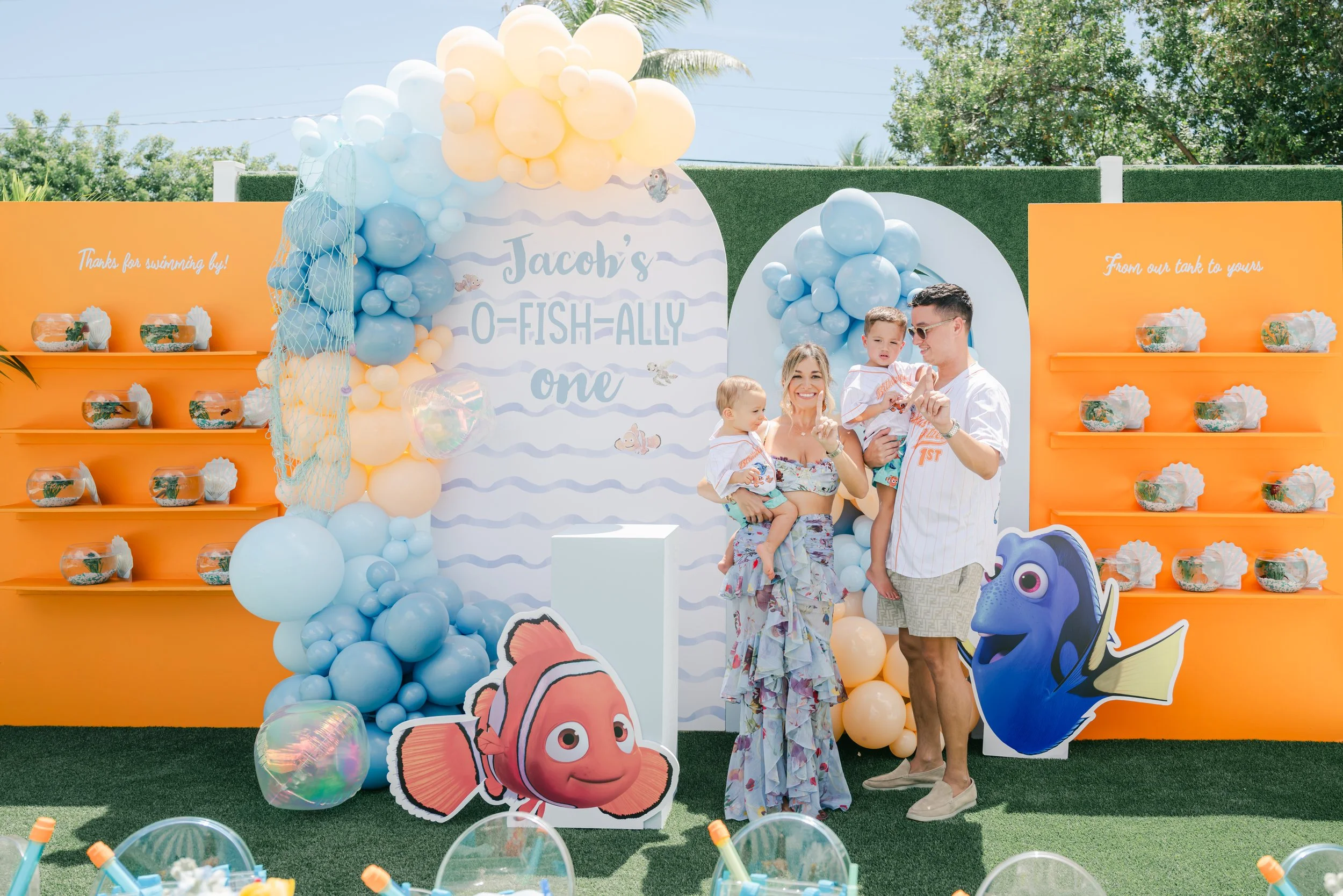 Family celebrating a child's first birthday with a Finding Nemo themed party. The backdrop features balloons, sea creatures, and ocean-themed decorations. A woman and man hold two young children, smiling and posing for the camera.