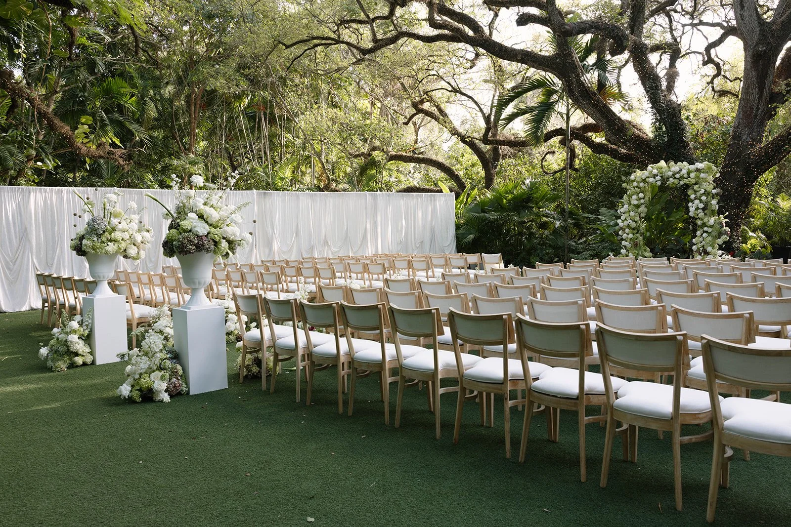 Outdoor wedding setup with white chairs, floral arrangements, and a white draped backdrop in a lush green garden.