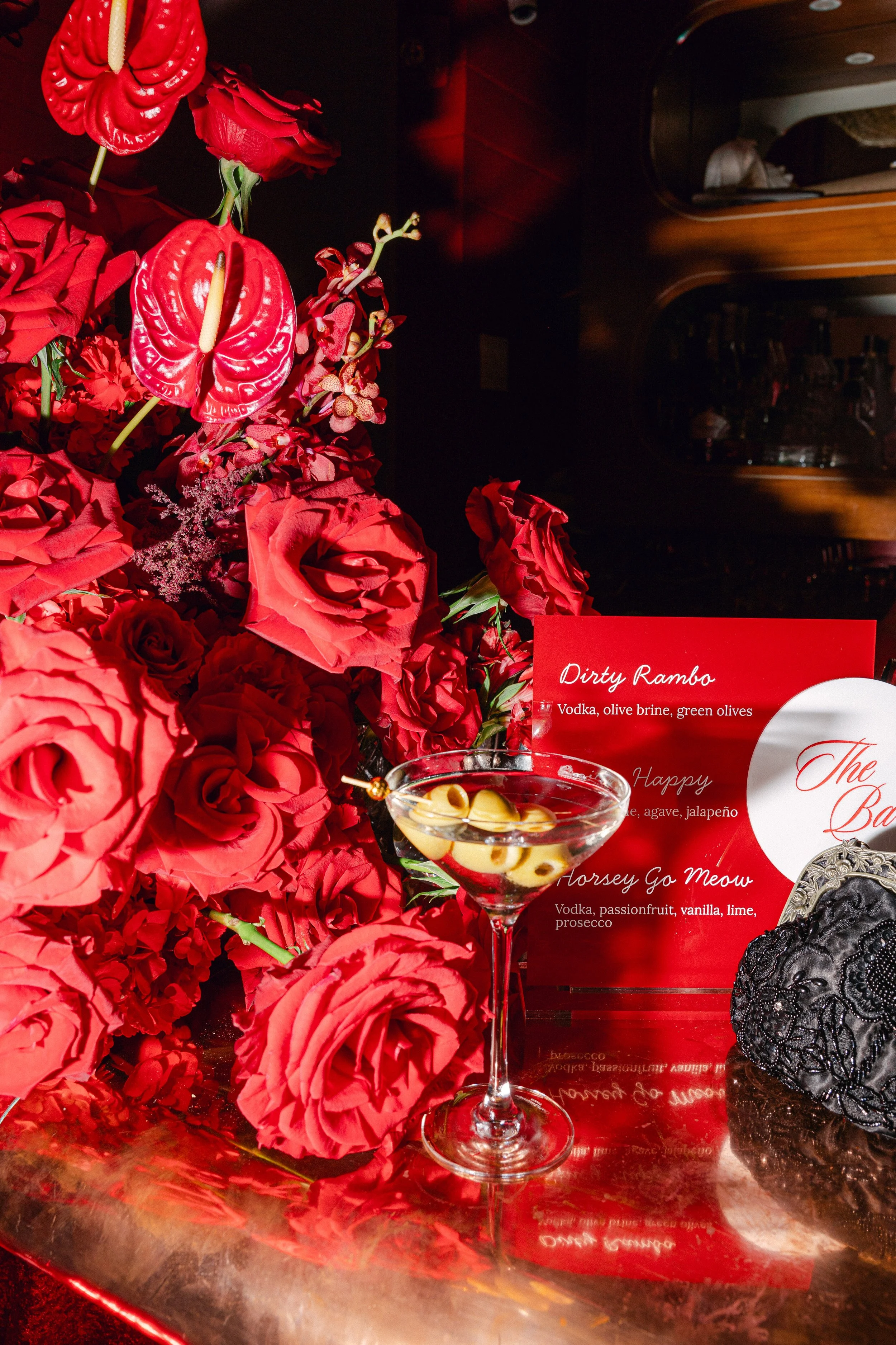 A bouquet of vibrant red flowers next to a bar menu displaying cocktails with alcohol, garnishes, and ingredients, along with a glass of cocktail with olives in a dimly-lit bar or restaurant setting.