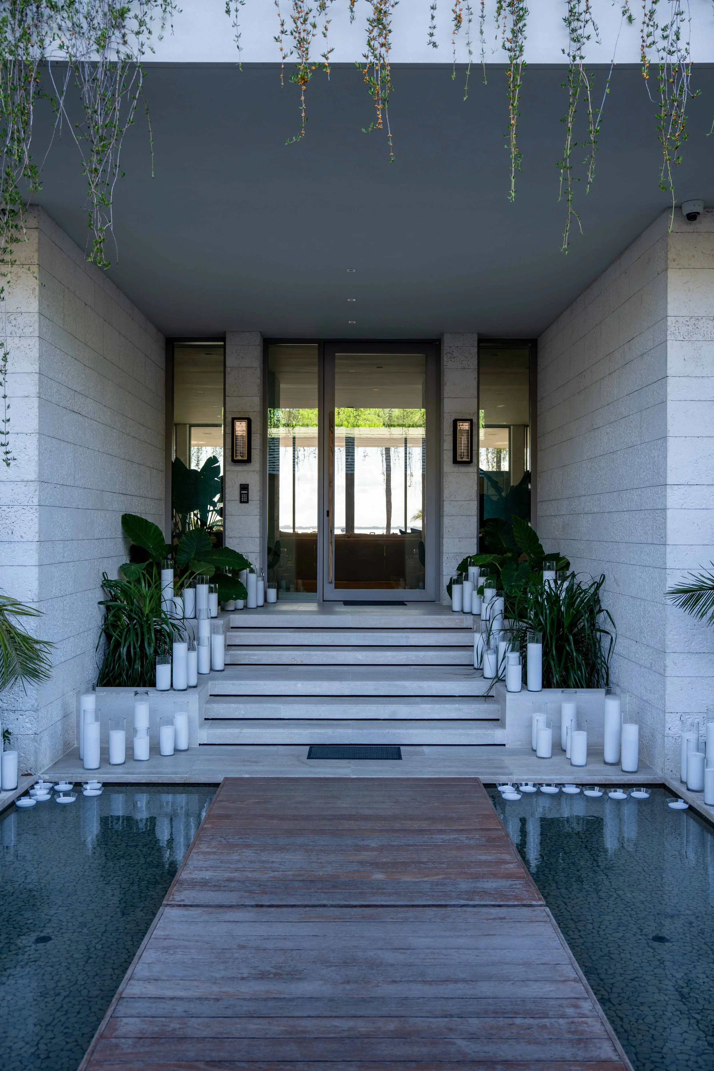 Front entrance of a modern building with a glass door, surrounded by green plants in white pots, and decorated with white candles on either side of the stairs and water at the bottom.