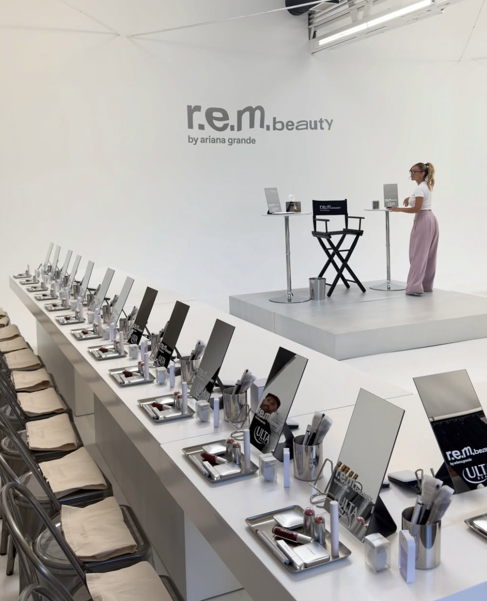 A beauty workshop setup with a long white table featuring multiple mirrors, hairbrushes, and makeup supplies, adjacent beige chairs, and a small stage with a station for a speaker or presenter on a raised platform.