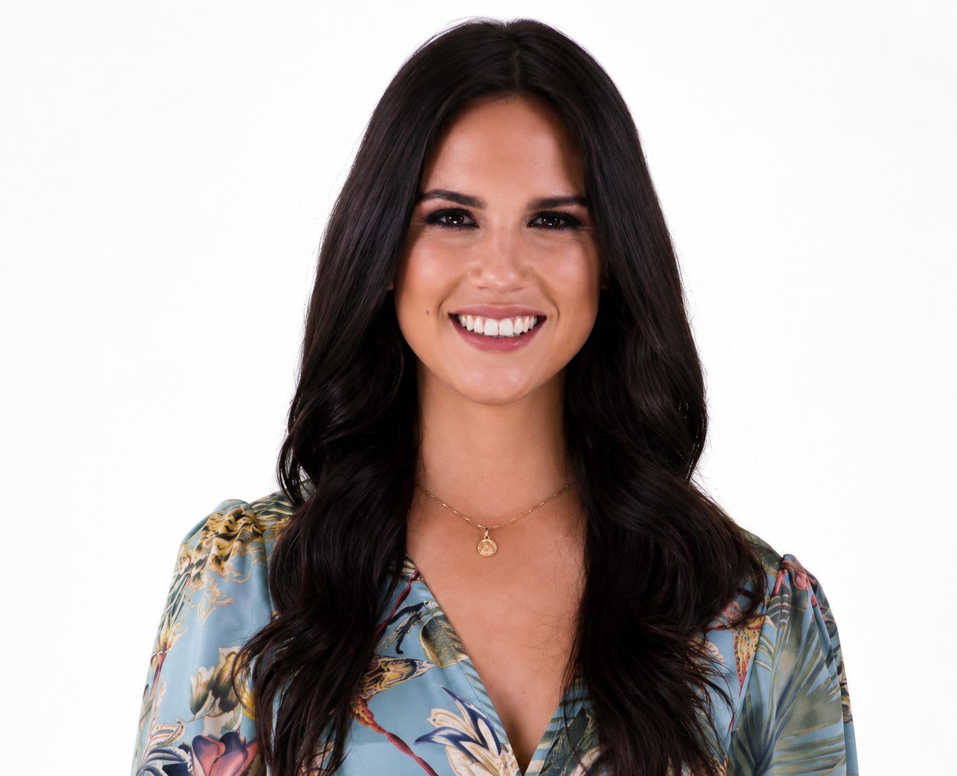 A woman with long dark wavy hair, wearing a floral blouse and a gold necklace, smiling against a plain white background.