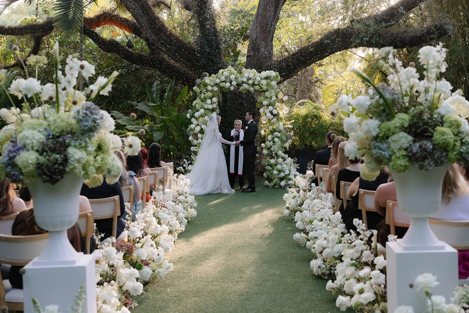 A wedding ceremony outdoors under a large tree with hanging lights. The bride and groom are holding hands and exchanging vows, with an officiant standing between them. The area is decorated with white flowers, and guests are seated on chairs facing t