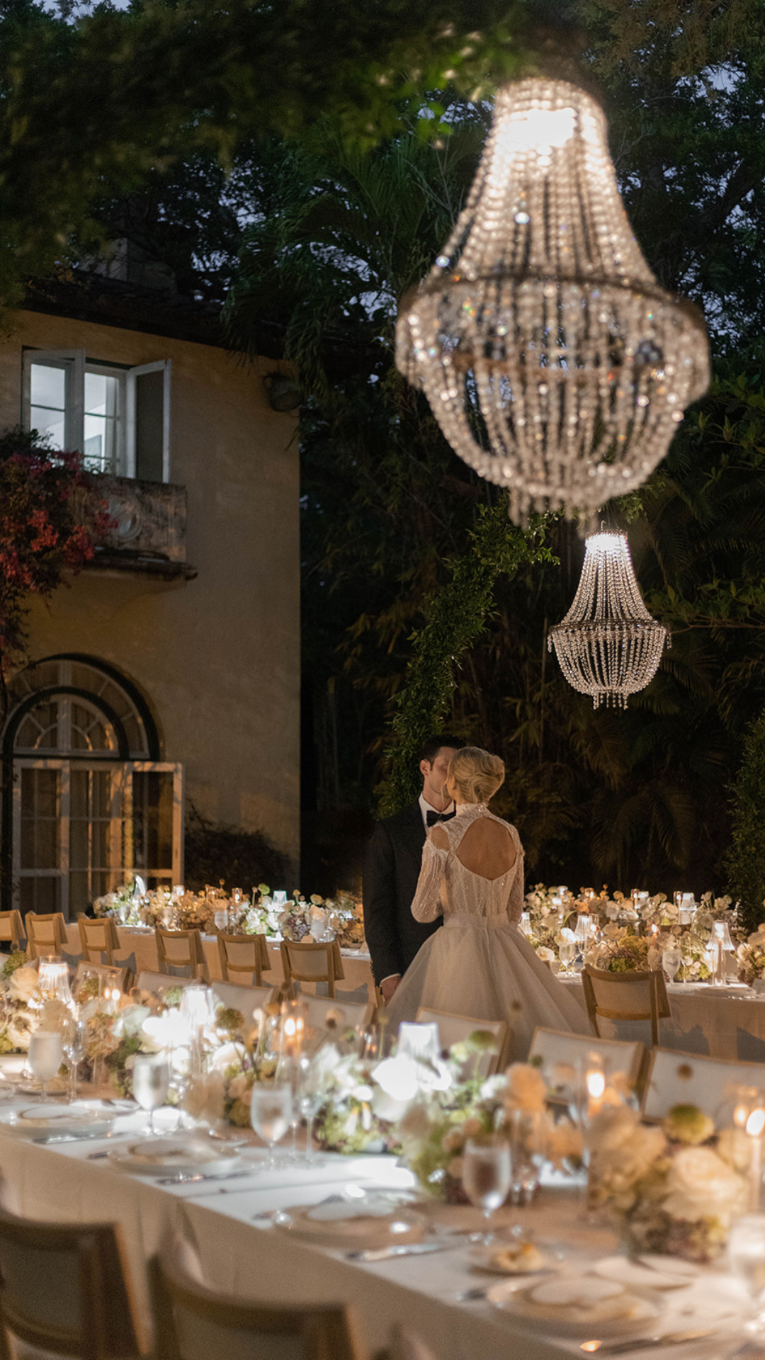 A bride and groom kiss at their wedding reception in an outdoor setting decorated with chandeliers and string lights. The scene features a long reception table with floral centerpieces, candlelit lamps, and elegant tableware, surrounded by greenery and a building with open windows.