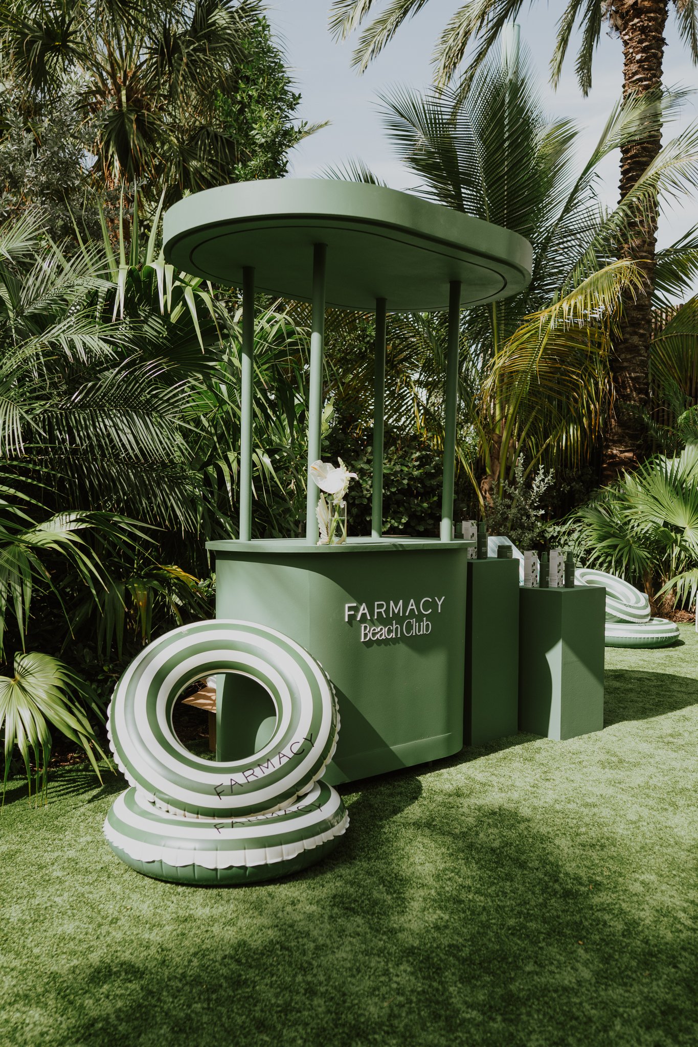 A green booth with the words "FARMACY Beach Club" at an outdoor location surrounded by lush tropical plants. There are white and green striped rings stacked in front of the booth.