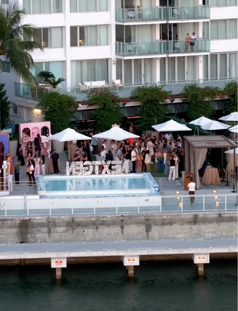 Outdoor event with people gathered under white umbrellas by the water, near a hotel or apartment building with balconies. There is a pool in the foreground, and decorative letters spelling 'BEXTEN' on the edge of the pool area. The scene appears to be a social gathering or party.
