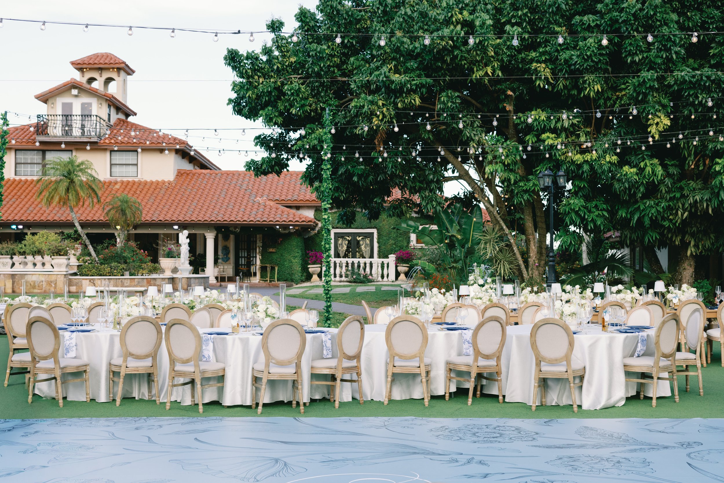 Outdoor event setup with round tables covered in white tablecloths, decorated with white floral centerpieces and set with glassware and plates. Surrounding the tables are beige chairs with oval backrests. In the background, there are large green tree