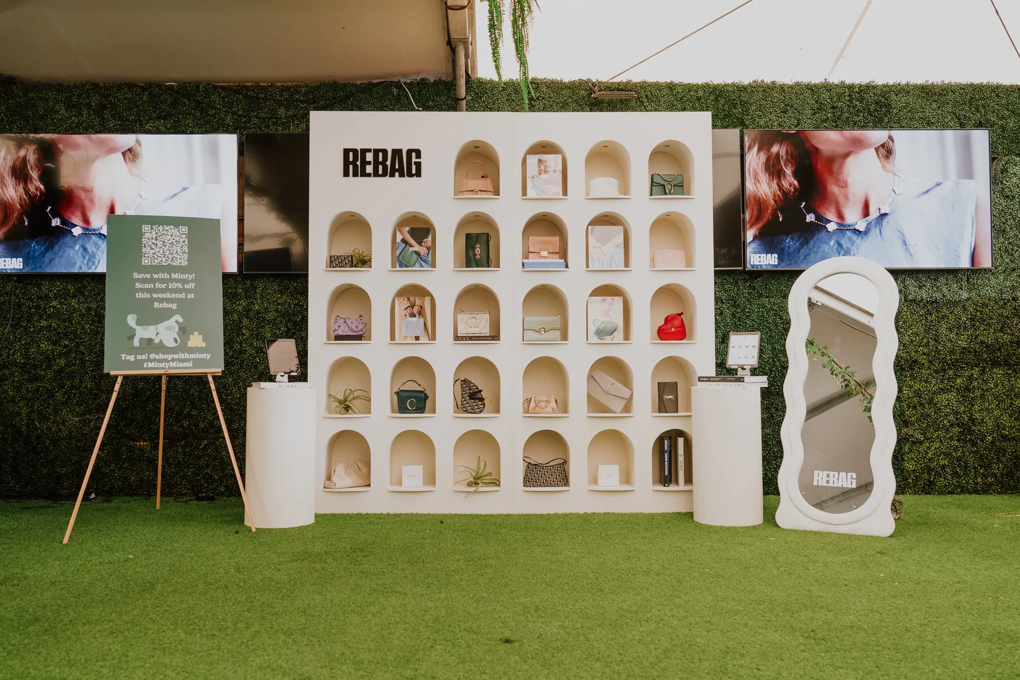 Display of handbags and accessories on a white shelf with multiple arched compartments at a store, with two large digital screens showing a woman wearing a necklace, and a QR code sign on a stand to the left.