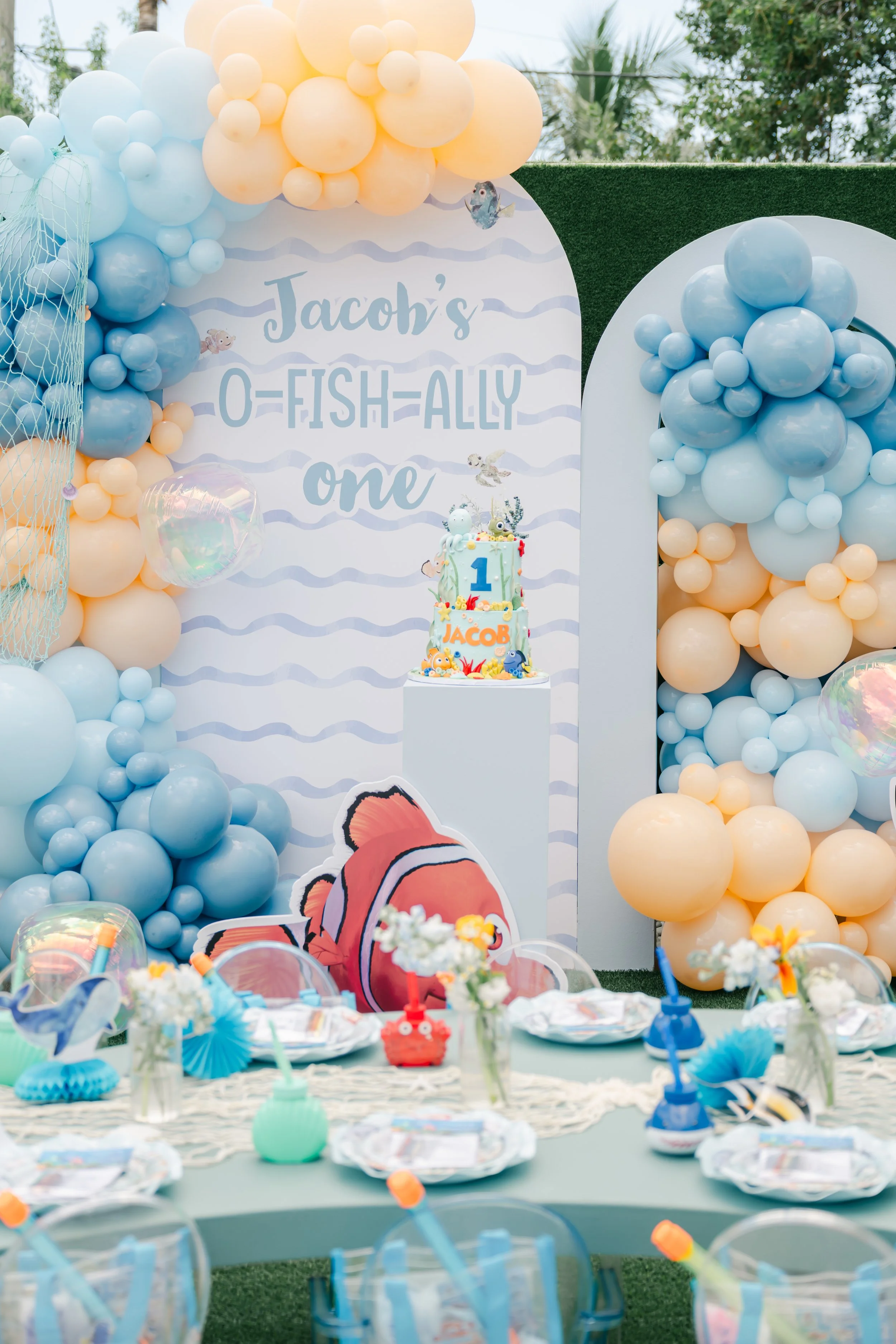 Underwater-themed birthday party setup with pastel-colored balloons, a decorated cake with an ocean theme, and party table with seashell, fish, and ocean decor.