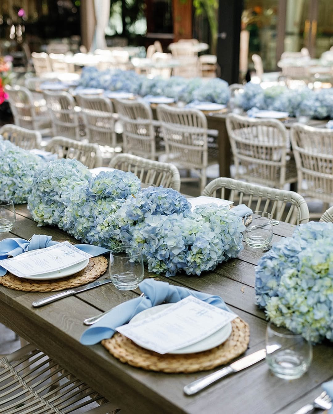 Long wooden table decorated with large blue and white hydrangea flower arrangements, place settings with woven placemats, white plates, blue cloth napkins, and clear glasses, set for an outdoor event or celebration.