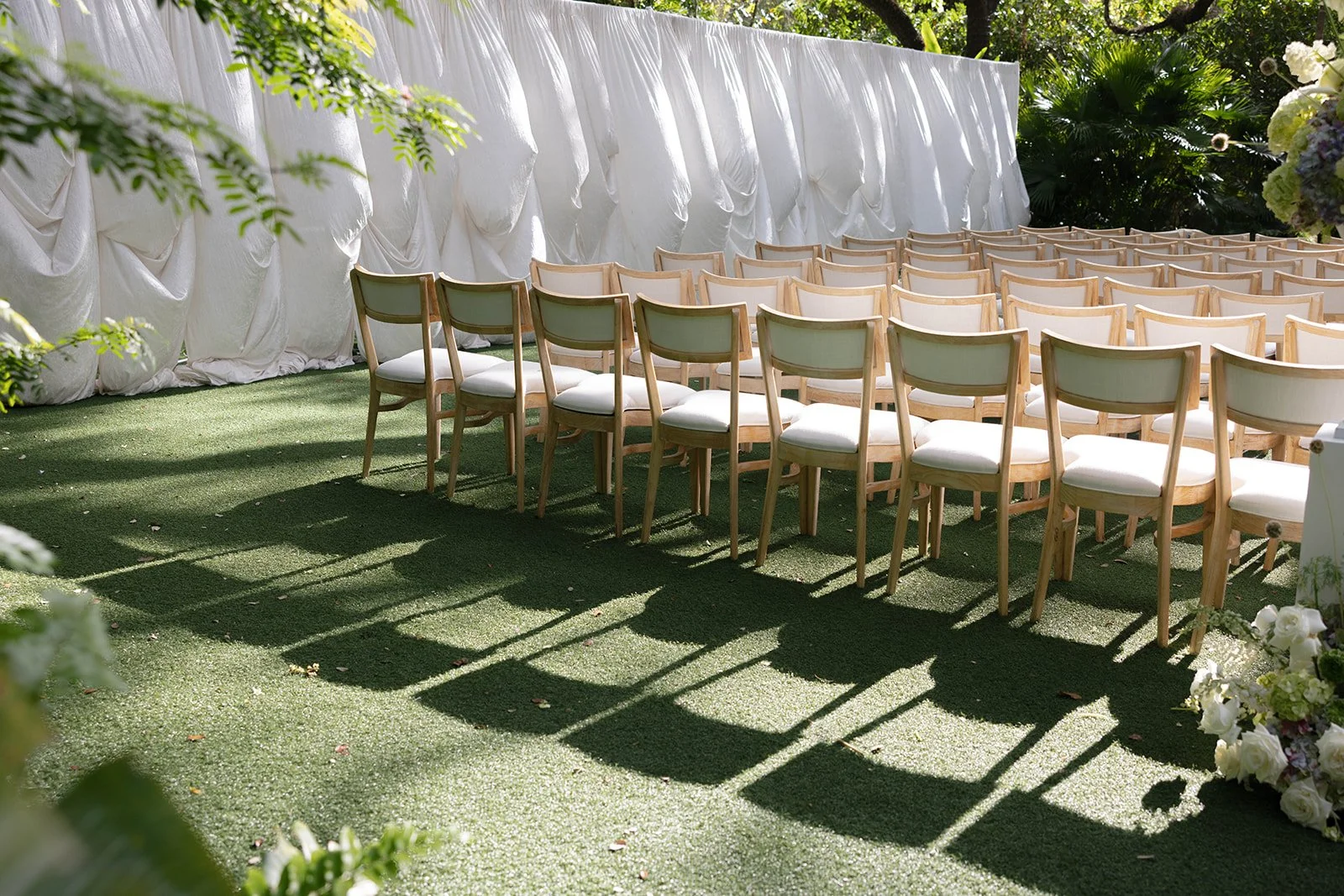 Outdoor wedding ceremony setup with rows of white and beige chairs on grassy ground, decorated with lush greenery and white flowers.