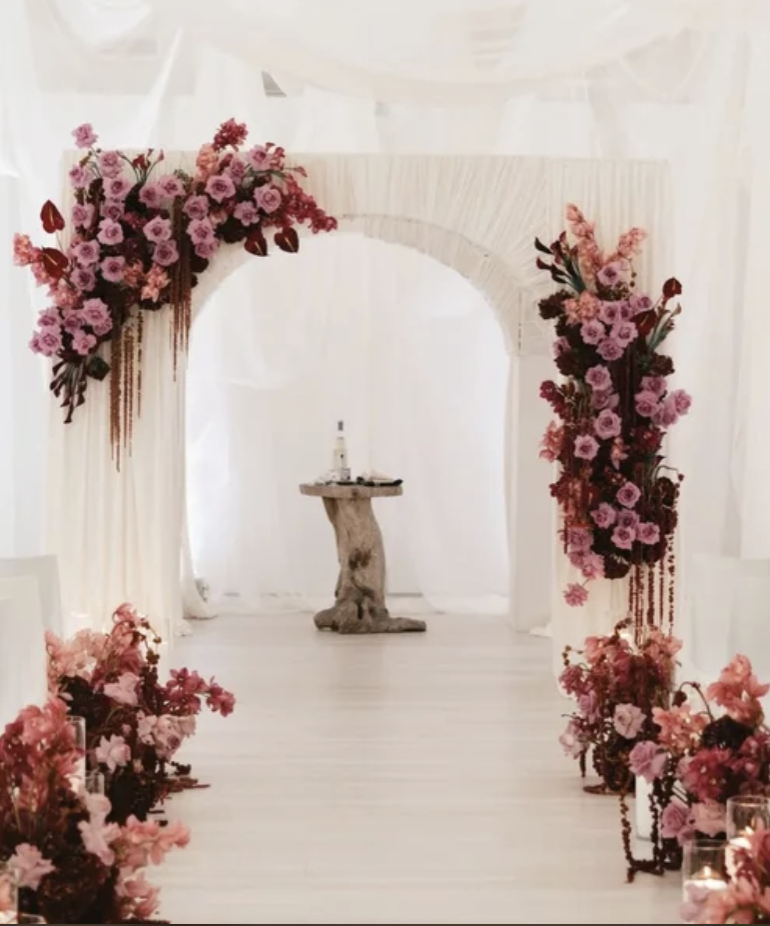 Flower-decorated wedding arch with pink and red flowers, white drapes, and a rustic wooden table in the background.